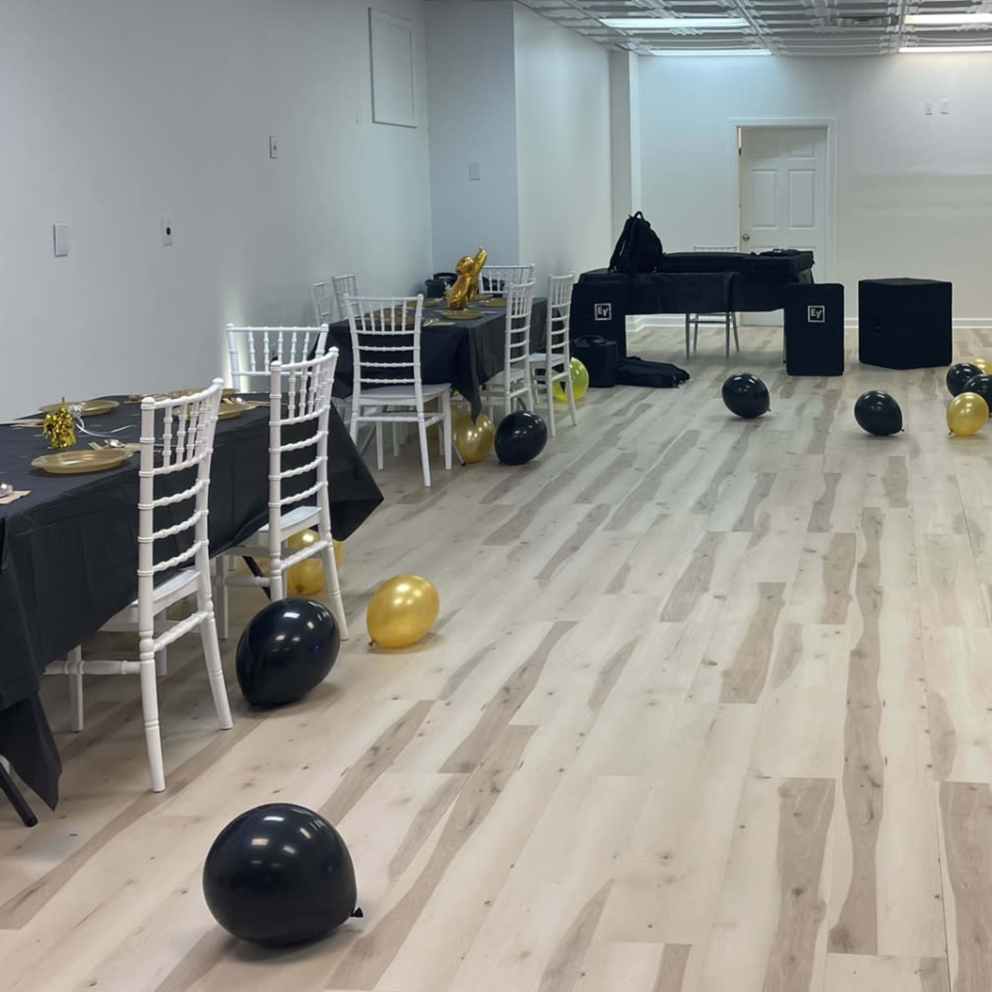 A spacious event room features tables set with black tablecloths, white chairs, and scattered black and gold balloons on the floor.