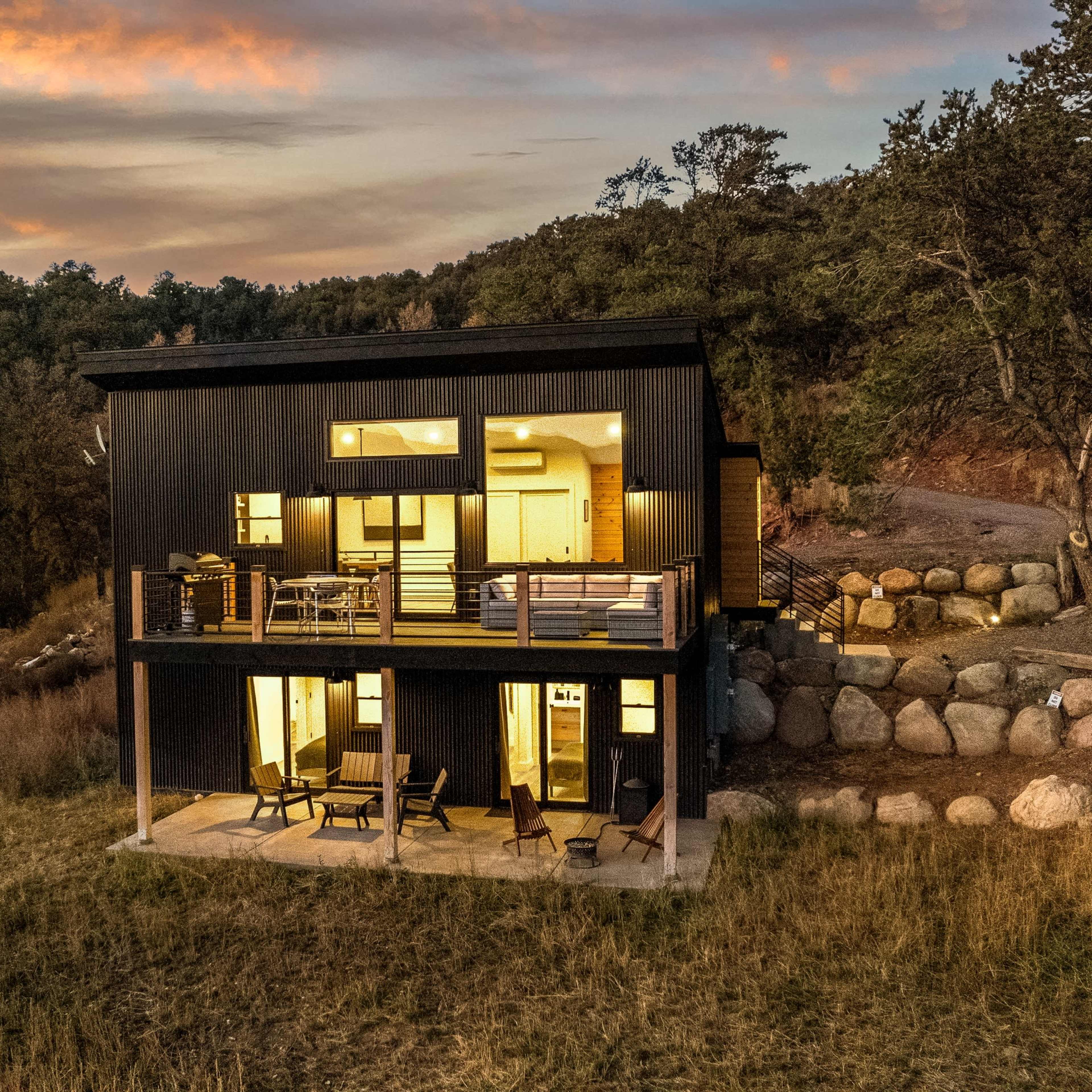 A modern black house with large windows is positioned on a hillside, surrounded by grass and boulders, as the sun sets in the background.