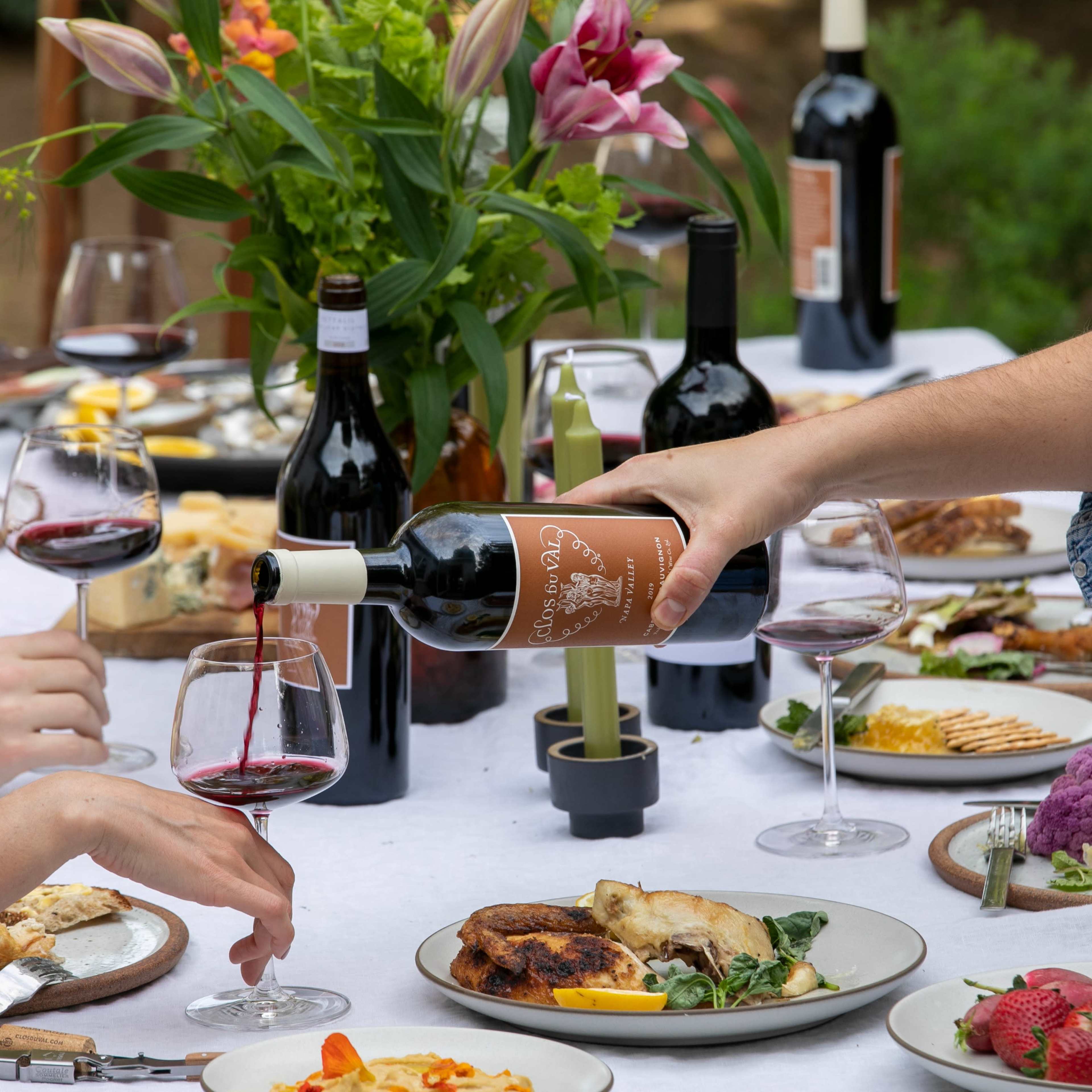 A person pours red wine into a glass at an outdoor dining table surrounded by various dishes and floral decorations.