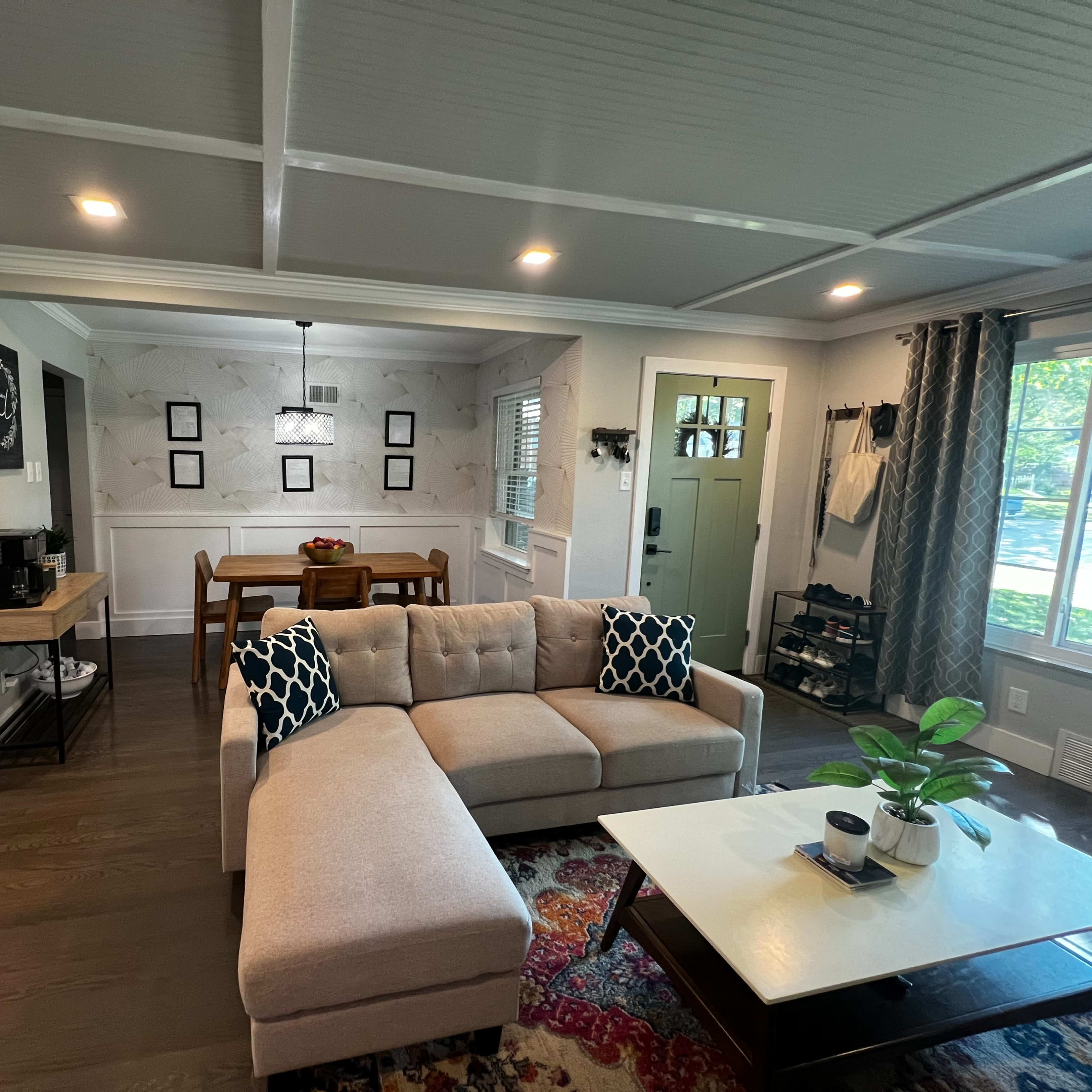 A spacious living room with a sectional sofa, a coffee table, and a dining area adjacent to a kitchen, illuminated by natural light from nearby windows.