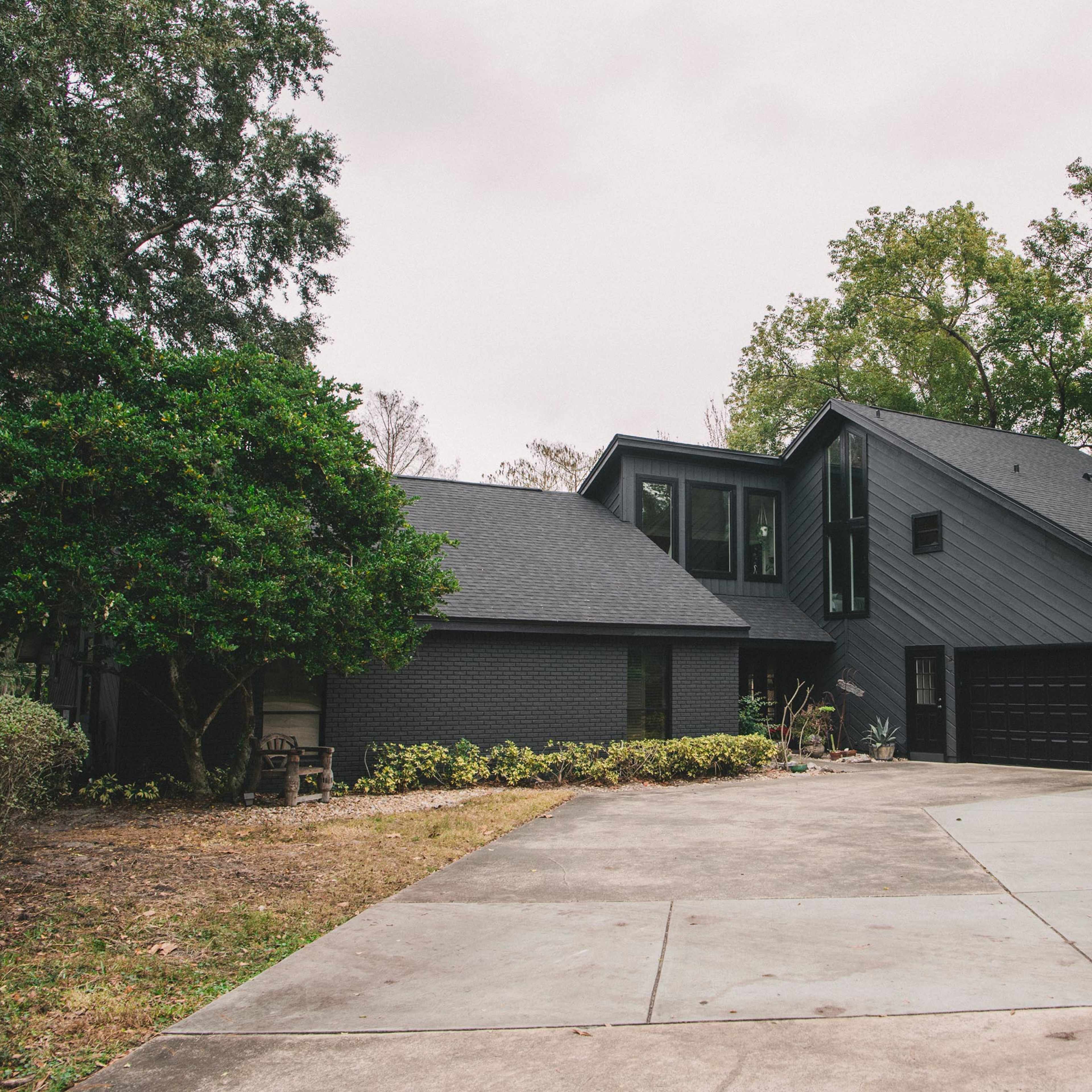 A modern black house with a sloped roof sits at the end of a curved driveway surrounded by greenery.
