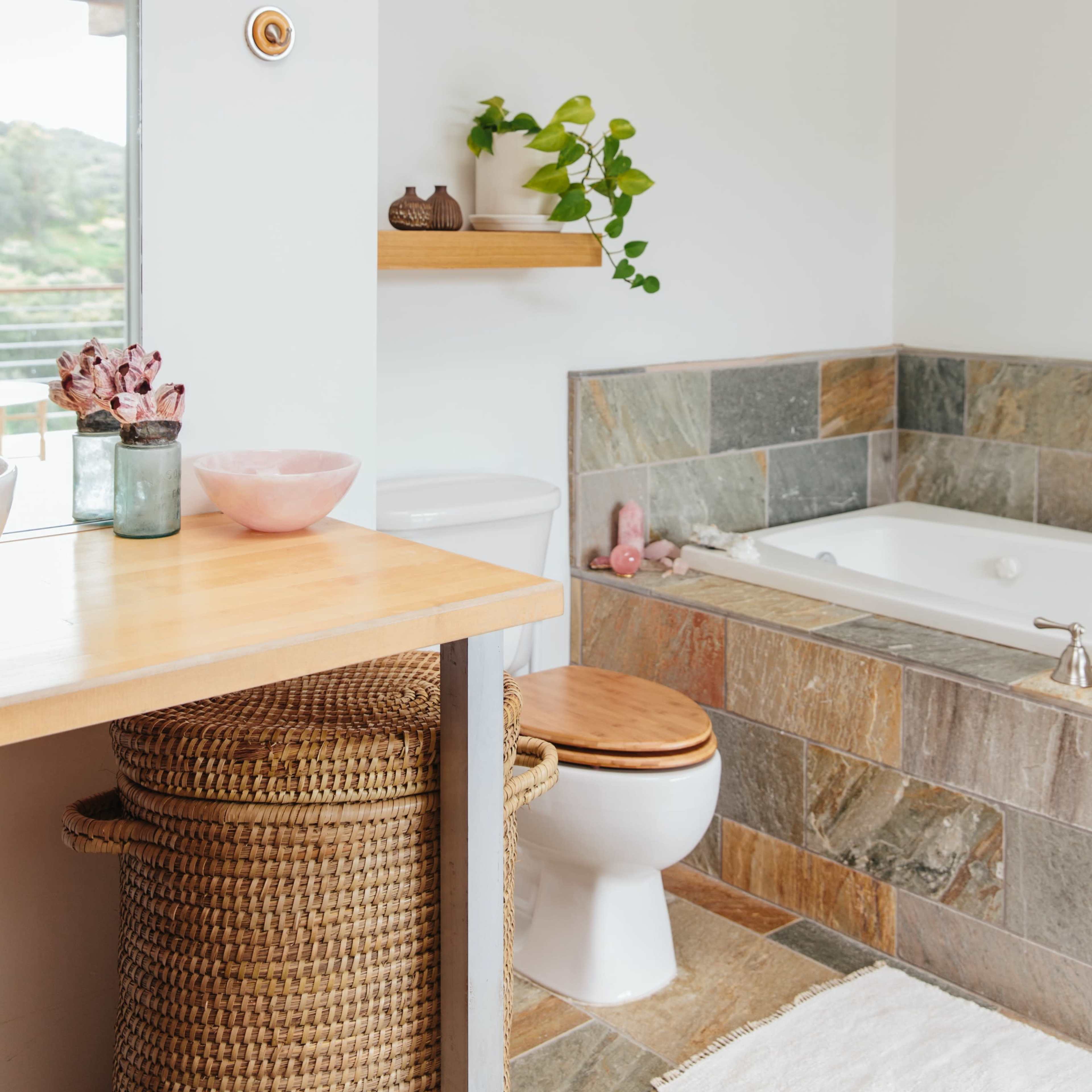 A modern bathroom features a bathtub with stone tiles, a toilet, a wooden shelf with plants, and a sink area with decorative items.