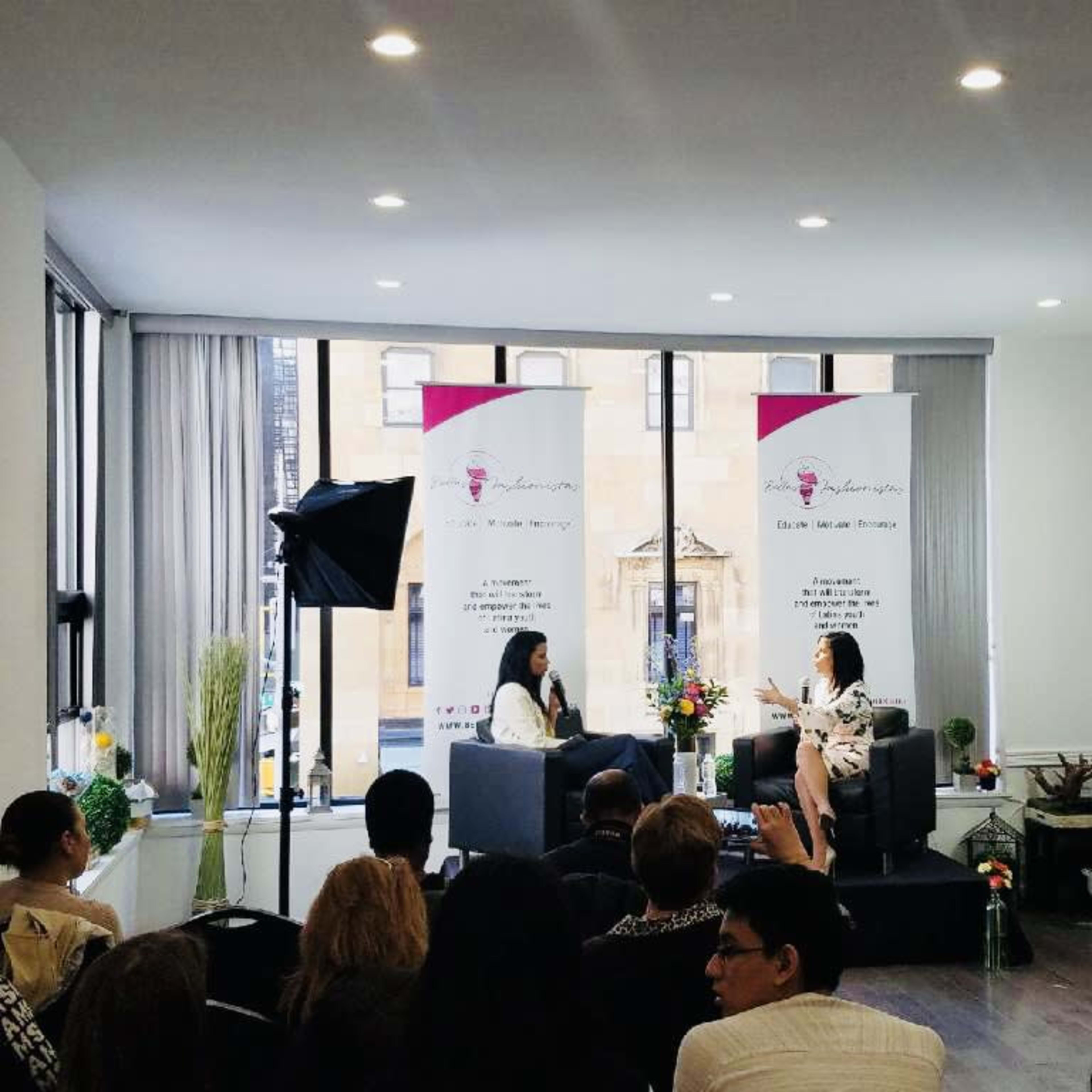 A woman interviews another in a well-lit room, surrounded by an audience and floral arrangements, with banners displayed in the background.