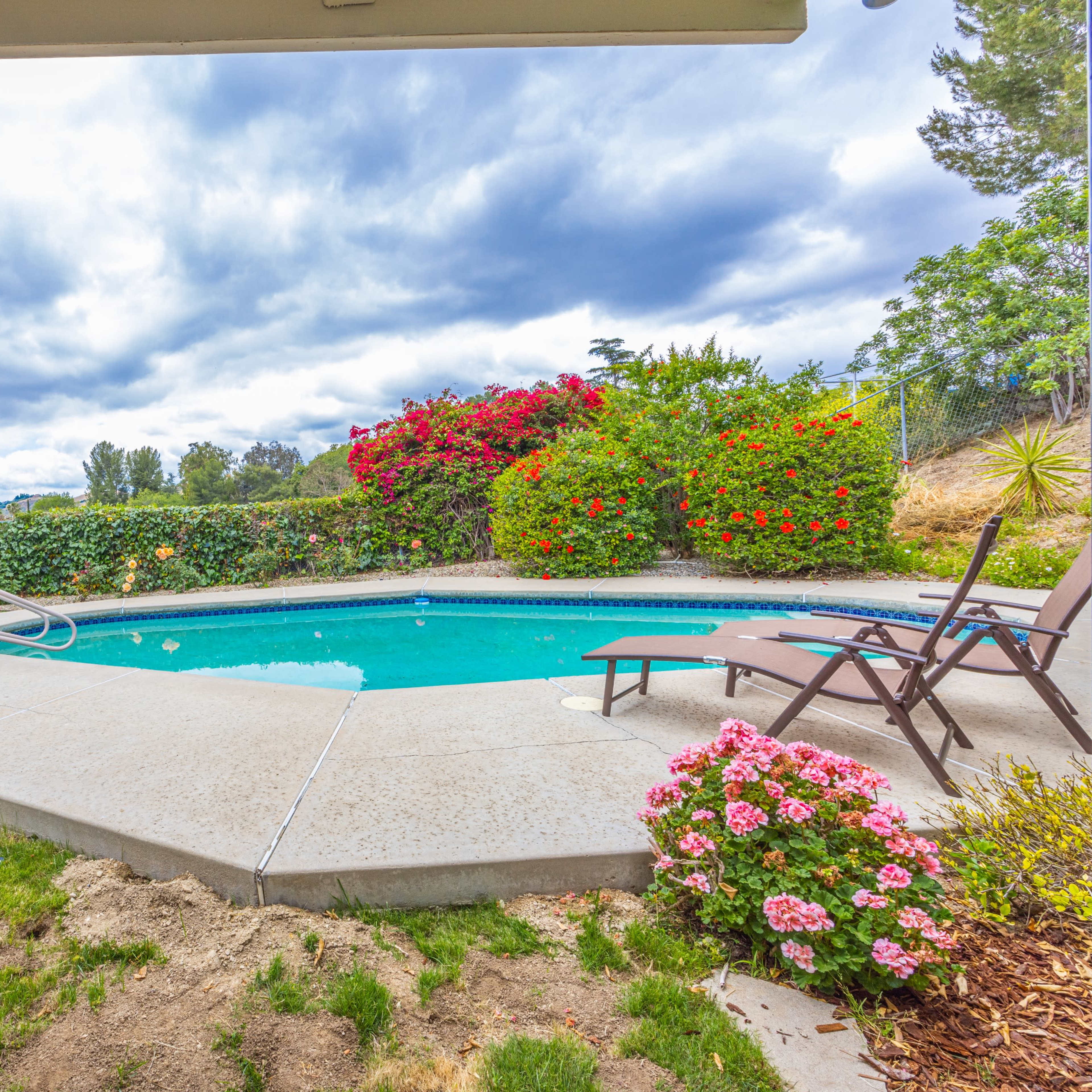 A pool area is surrounded by manicured grass and flowering shrubs under a cloudy sky.