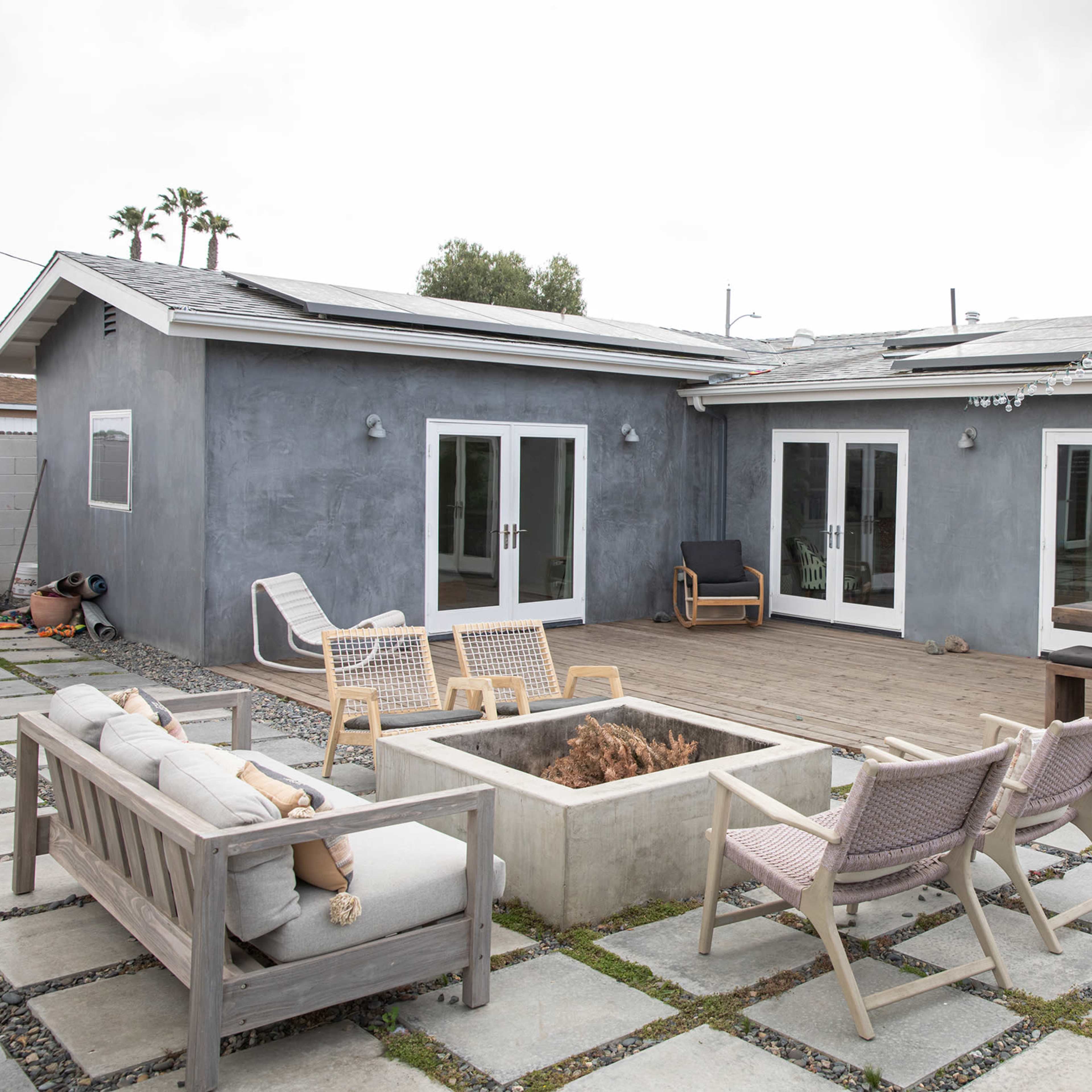 A modern patio area featuring a fire pit surrounded by outdoor seating, with a gray house in the background and string lights overhead.
