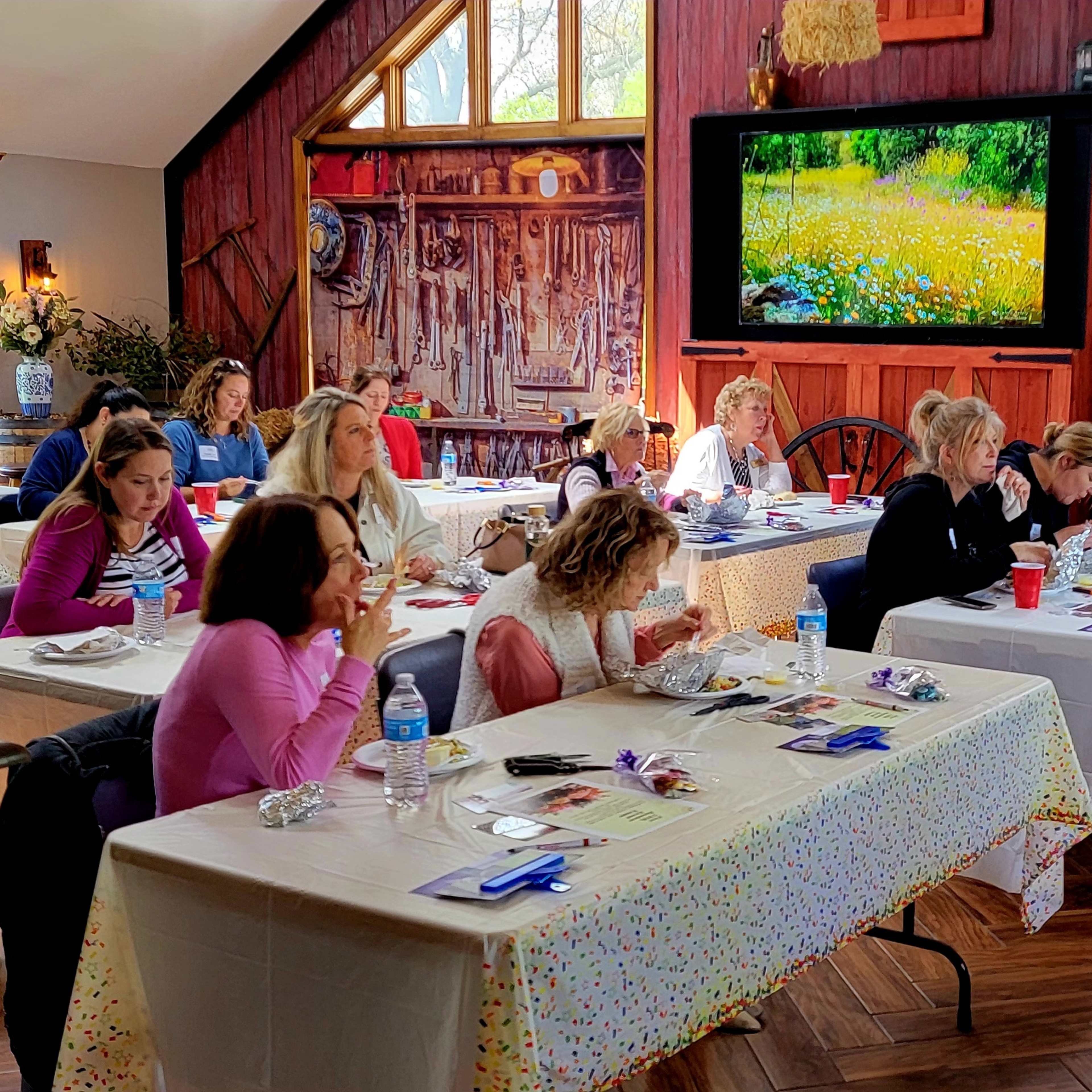 A group of women sits at long tables in a rustic room decorated with farm-themed elements, engaged in a dining or workshop activity.