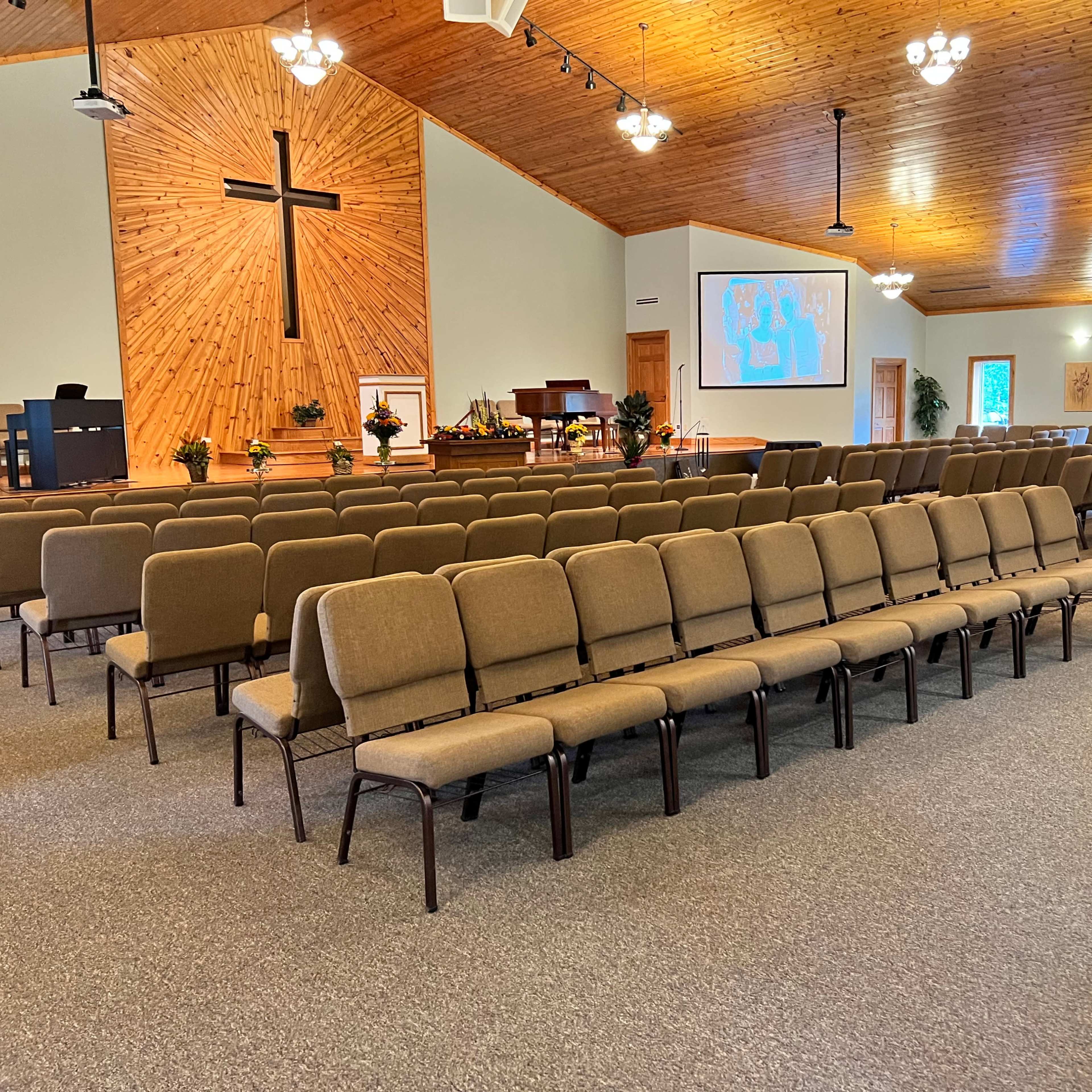 A spacious church interior with rows of neatly arranged chairs, a wooden cross on the wall, and a large projector screen displaying a map.