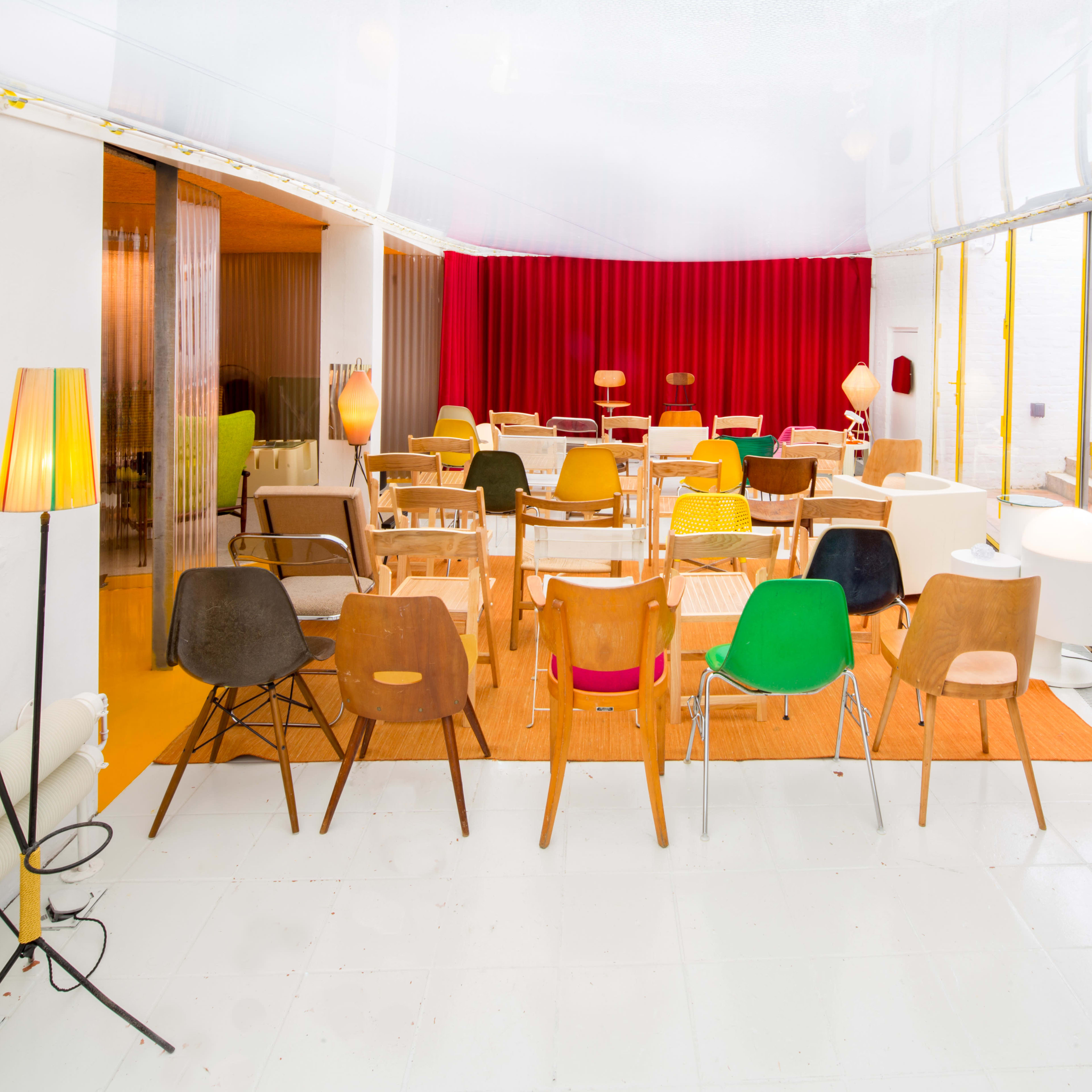 A brightly lit room with various chairs arranged in rows facing a red curtain, with decorative lamps and plants present in the space.