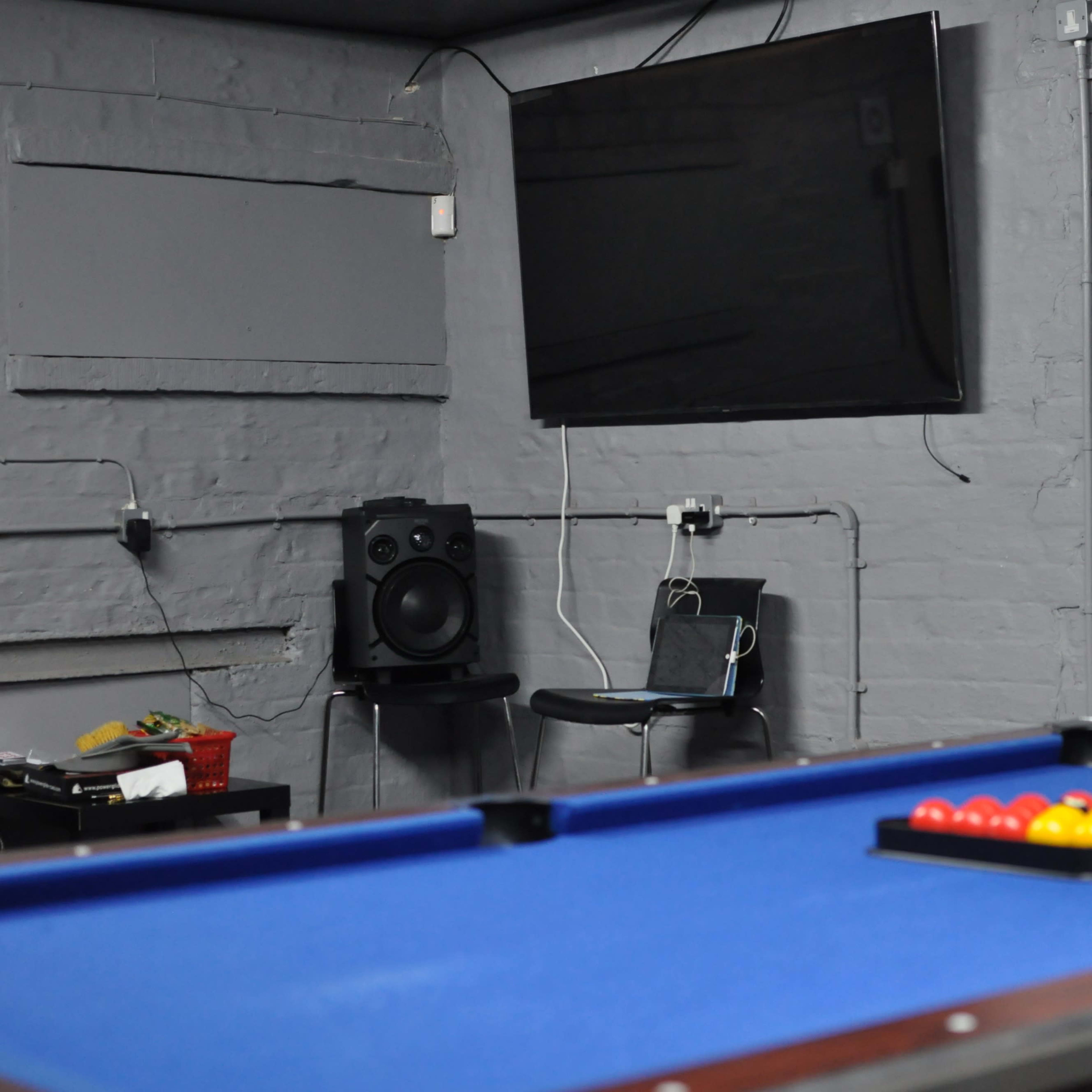 A blue pool table is set up with a rack of colorful balls in a dimly lit room featuring a flat-screen TV and a doorway.