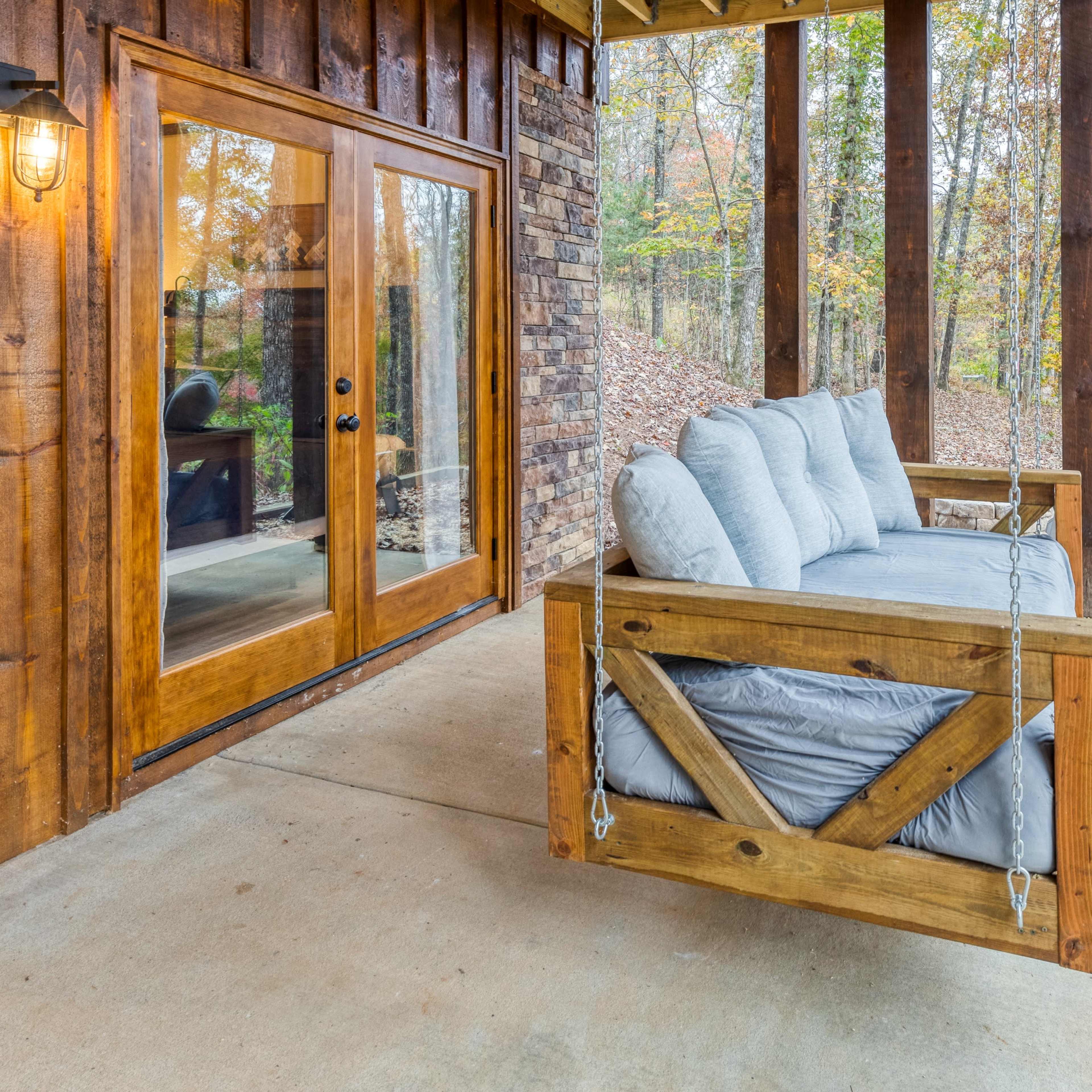 A wooden porch features a suspended bench and large windows overlooking a forested area.