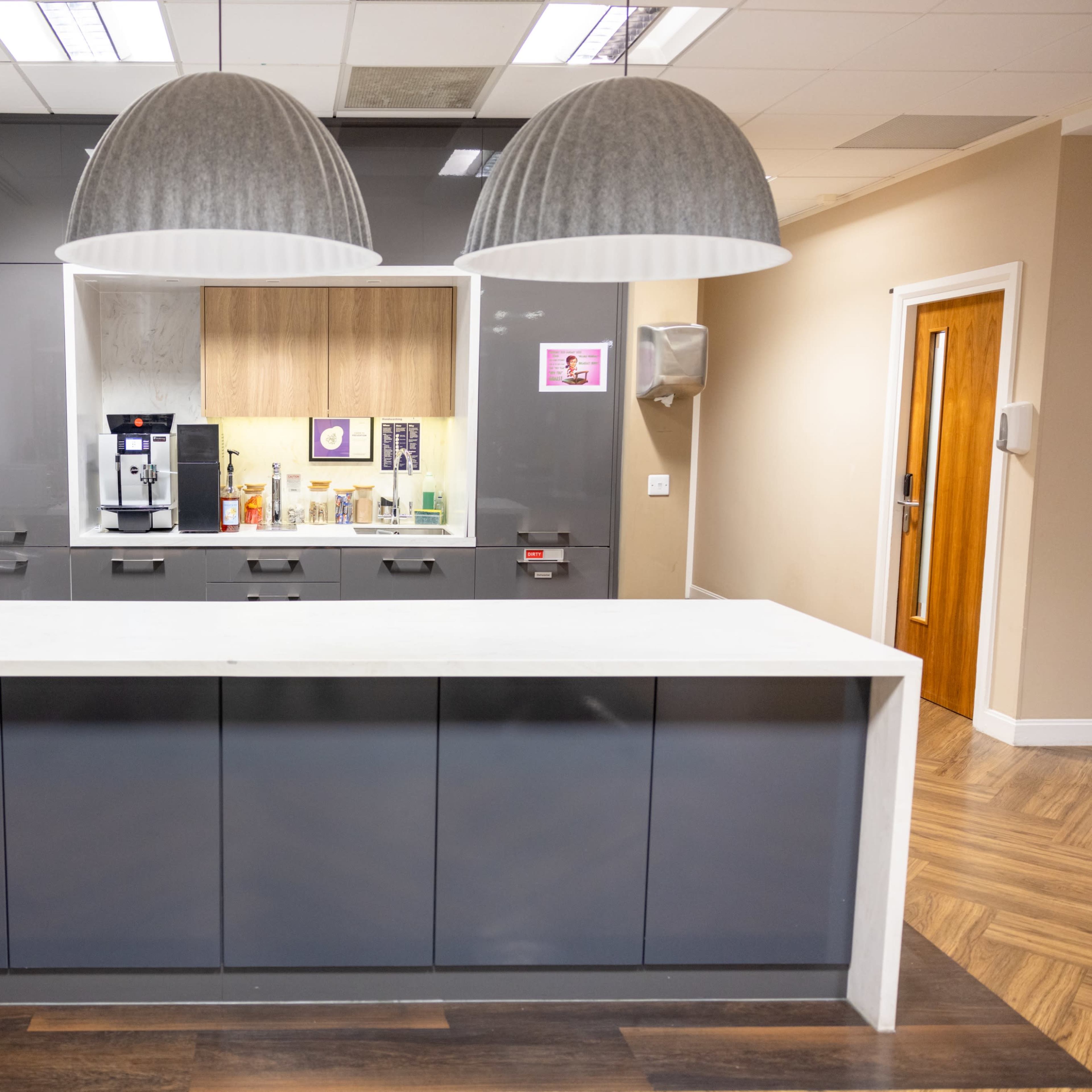 A modern kitchen area with a sleek gray countertop, three large pendant lights, and appliances including a coffee machine and microwaves.