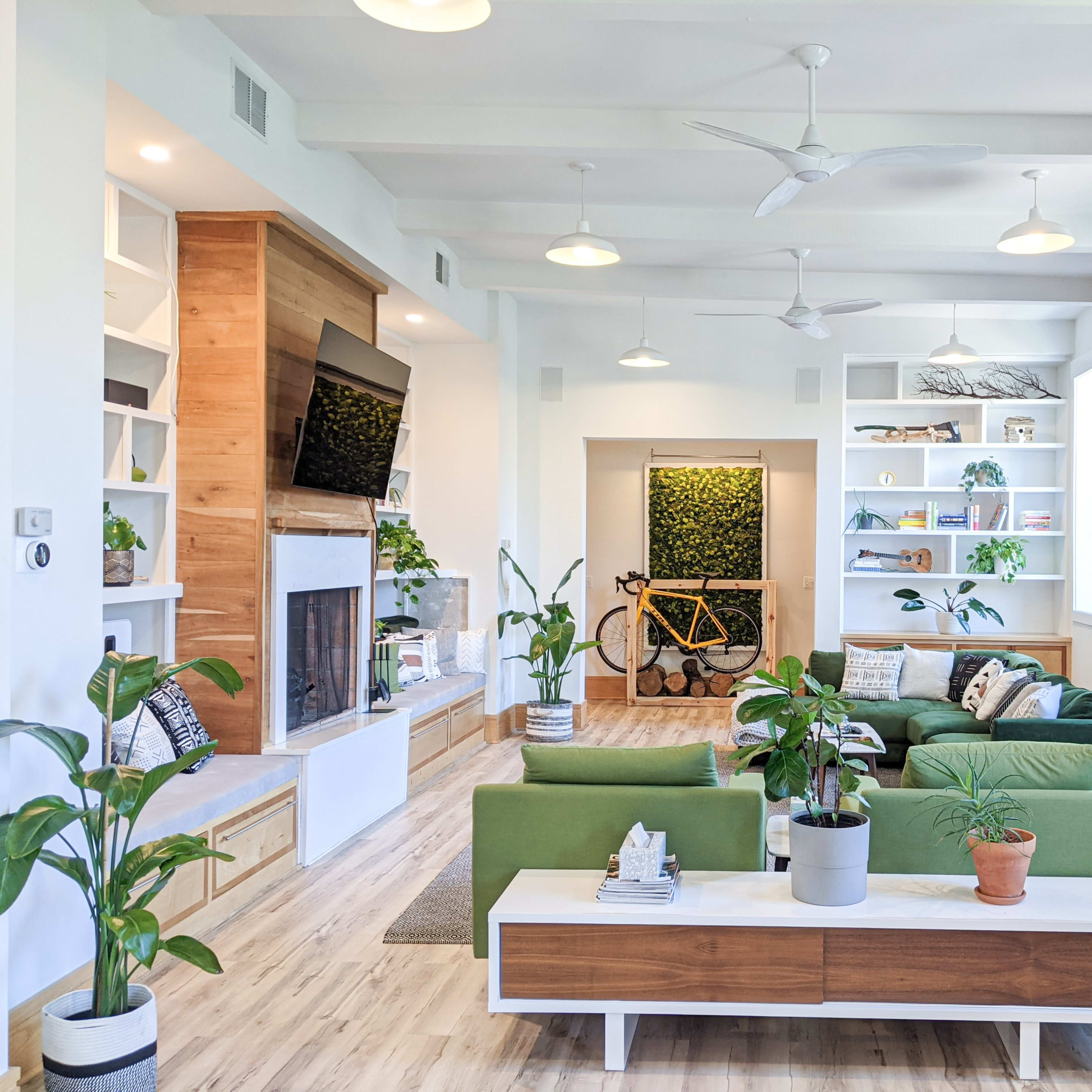 The living room features green sofas, a wooden coffee table, a fireplace, shelves filled with plants and decor, and a bicycle leaning against a wall.