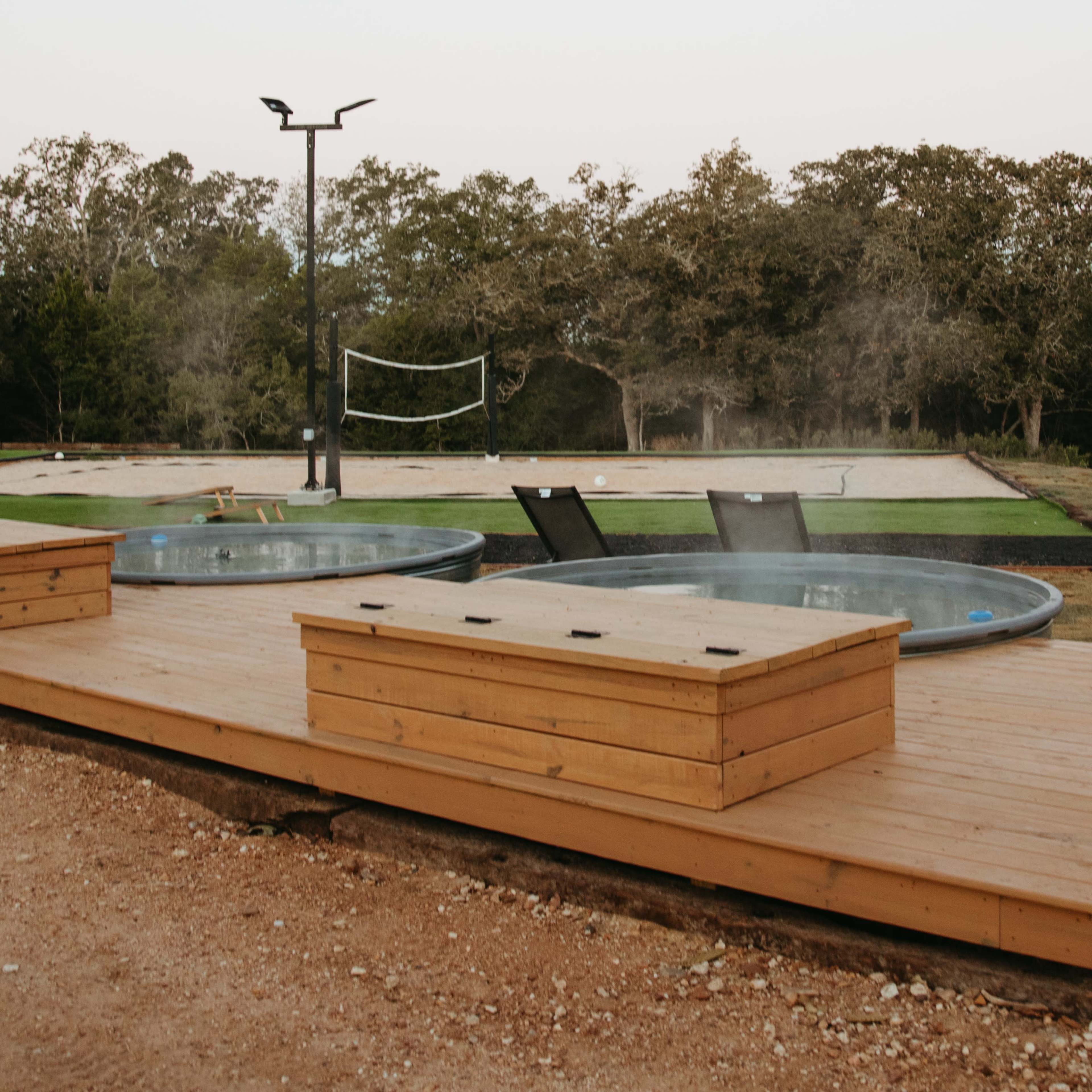 Two hot tubs situated on a wooden deck with a volleyball net and lawn in the background.