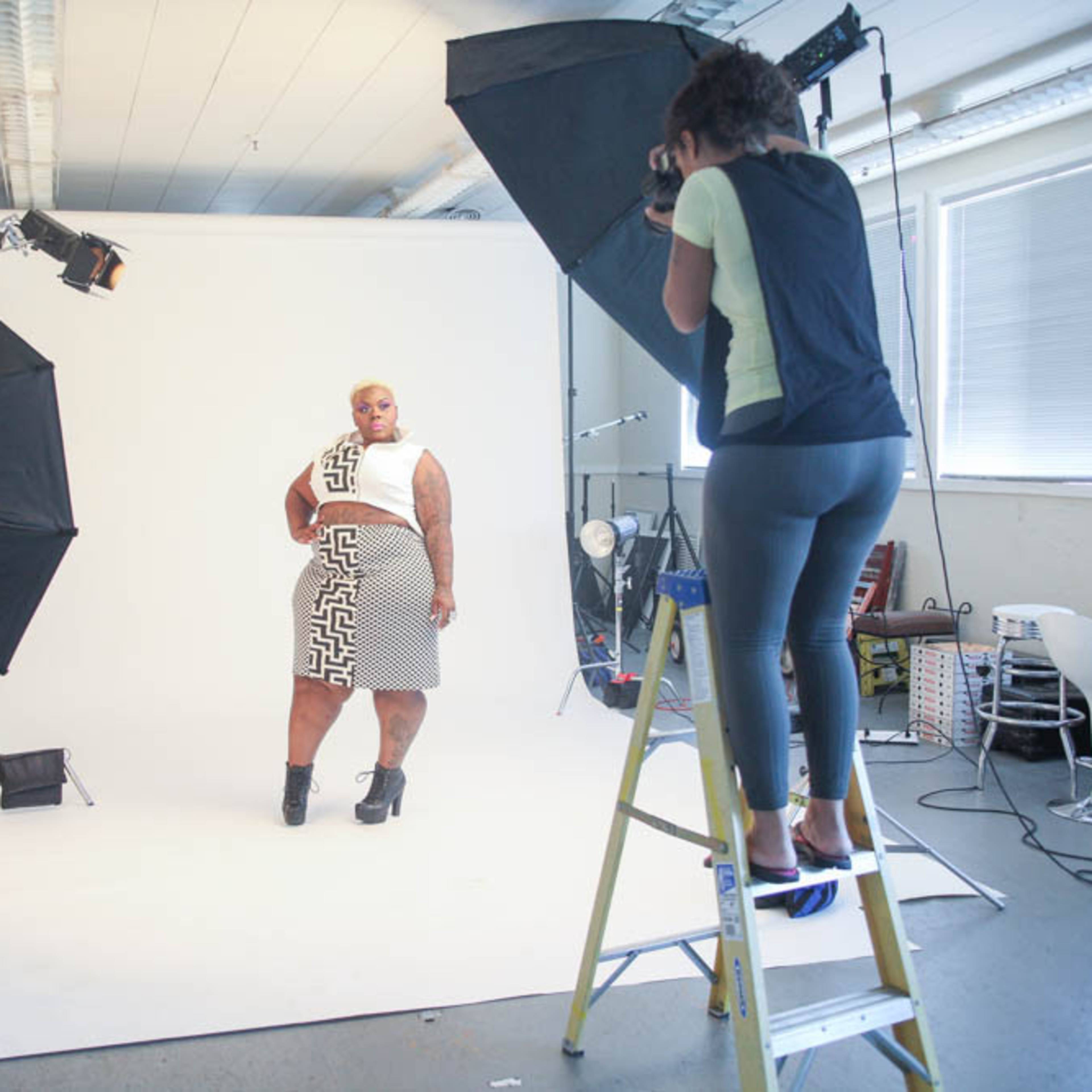 A photographer captures a model posing in front of a large white backdrop in a studio, with studio lights set up around them.