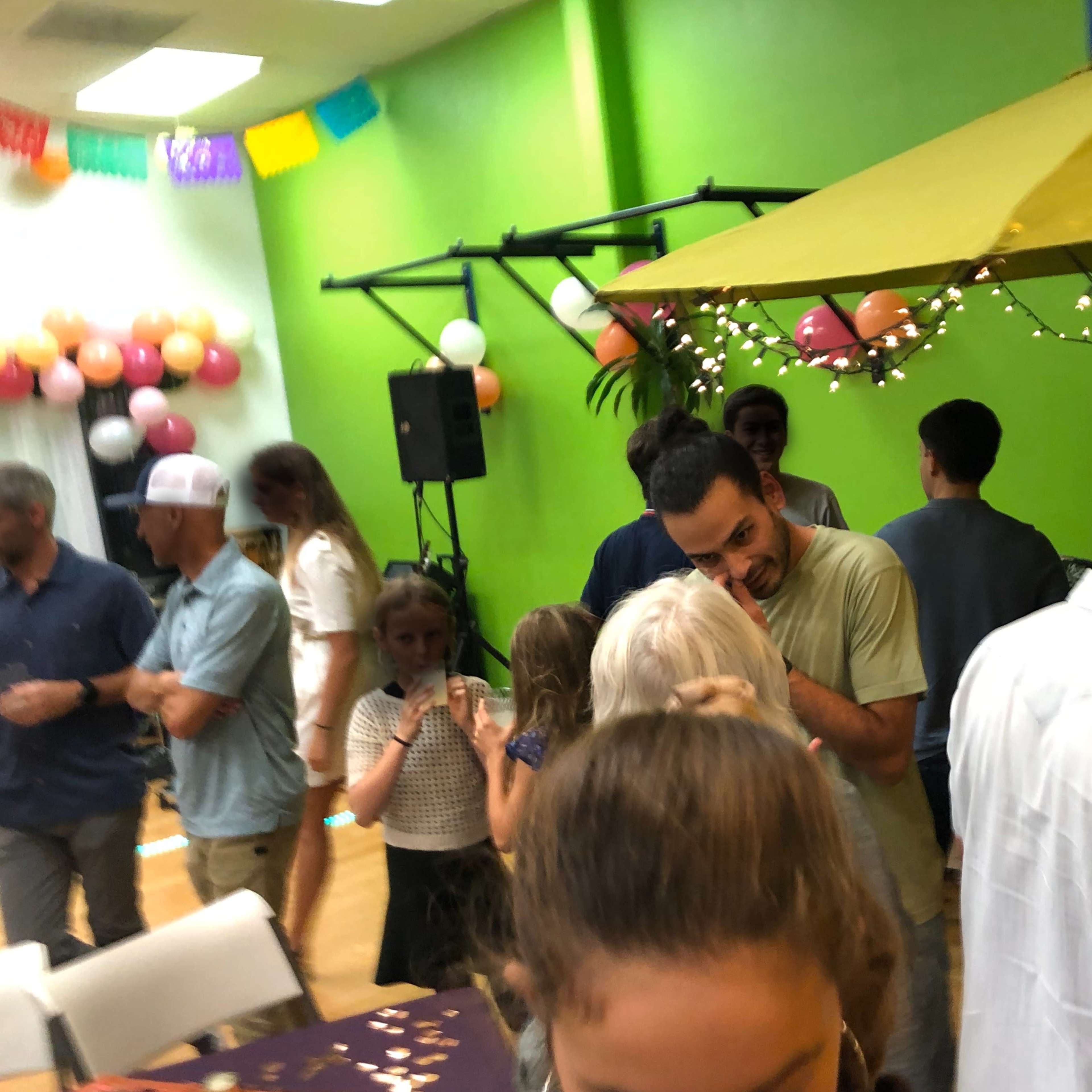 A group of people gather in a decorated room with green walls, socializing around a yellow canopy and string lights.