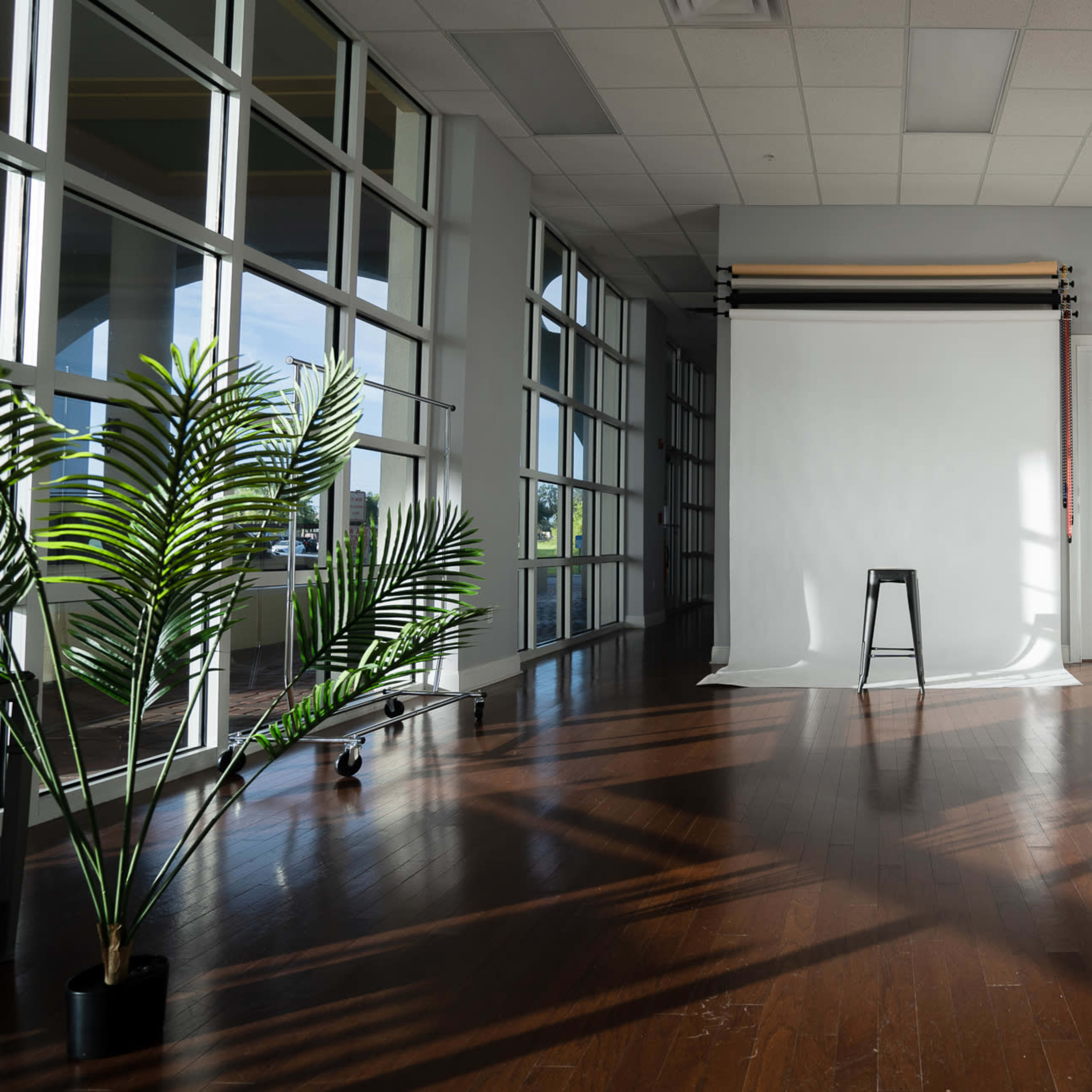 A spacious studio with a white backdrop, a black stool in the center, and colorful vertical stripes against a wall, illuminated by natural light from large windows.