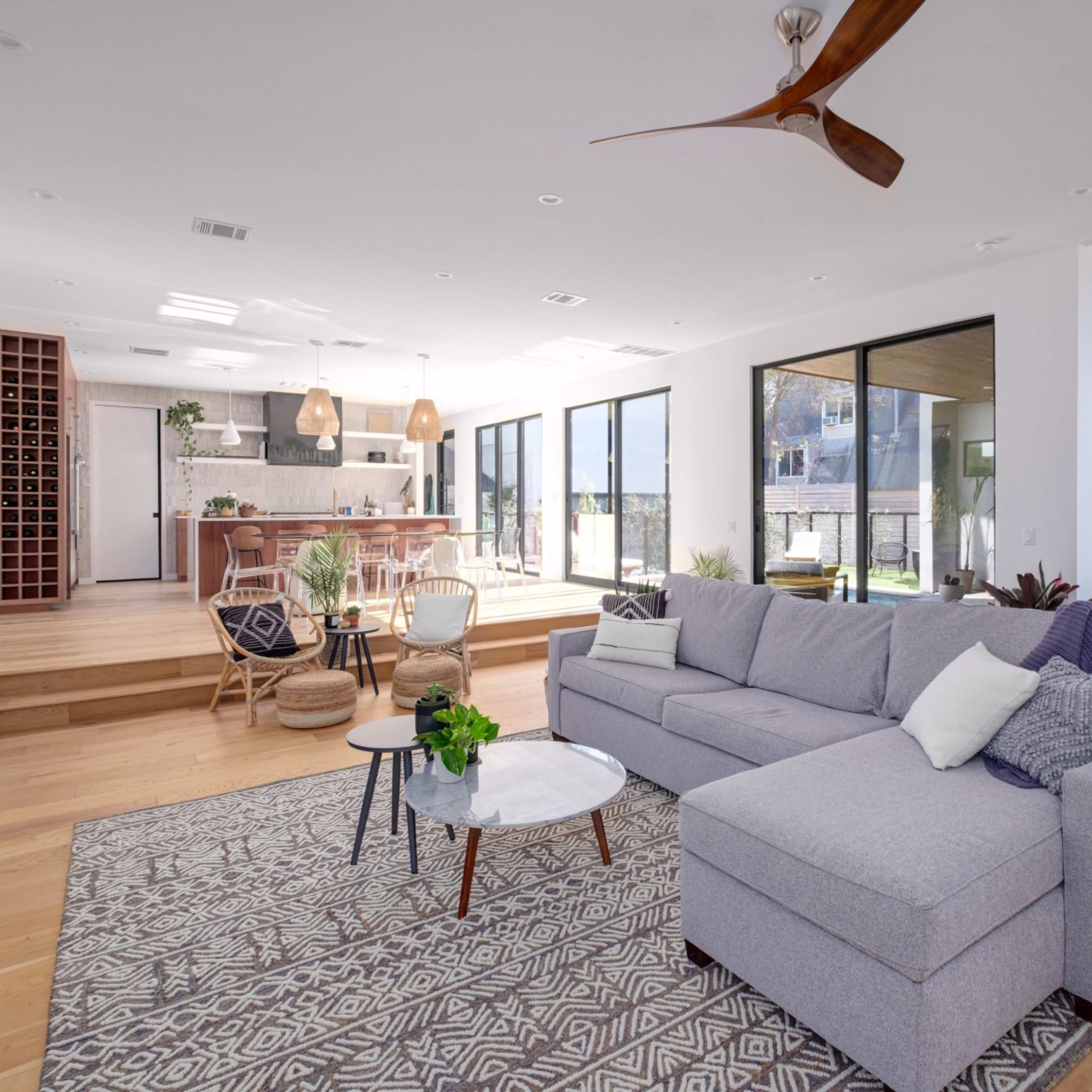 A modern living room with a gray sectional sofa, a round white coffee table, and large windows that open to an outdoor area, complemented by wooden flooring and a staircase in the background.