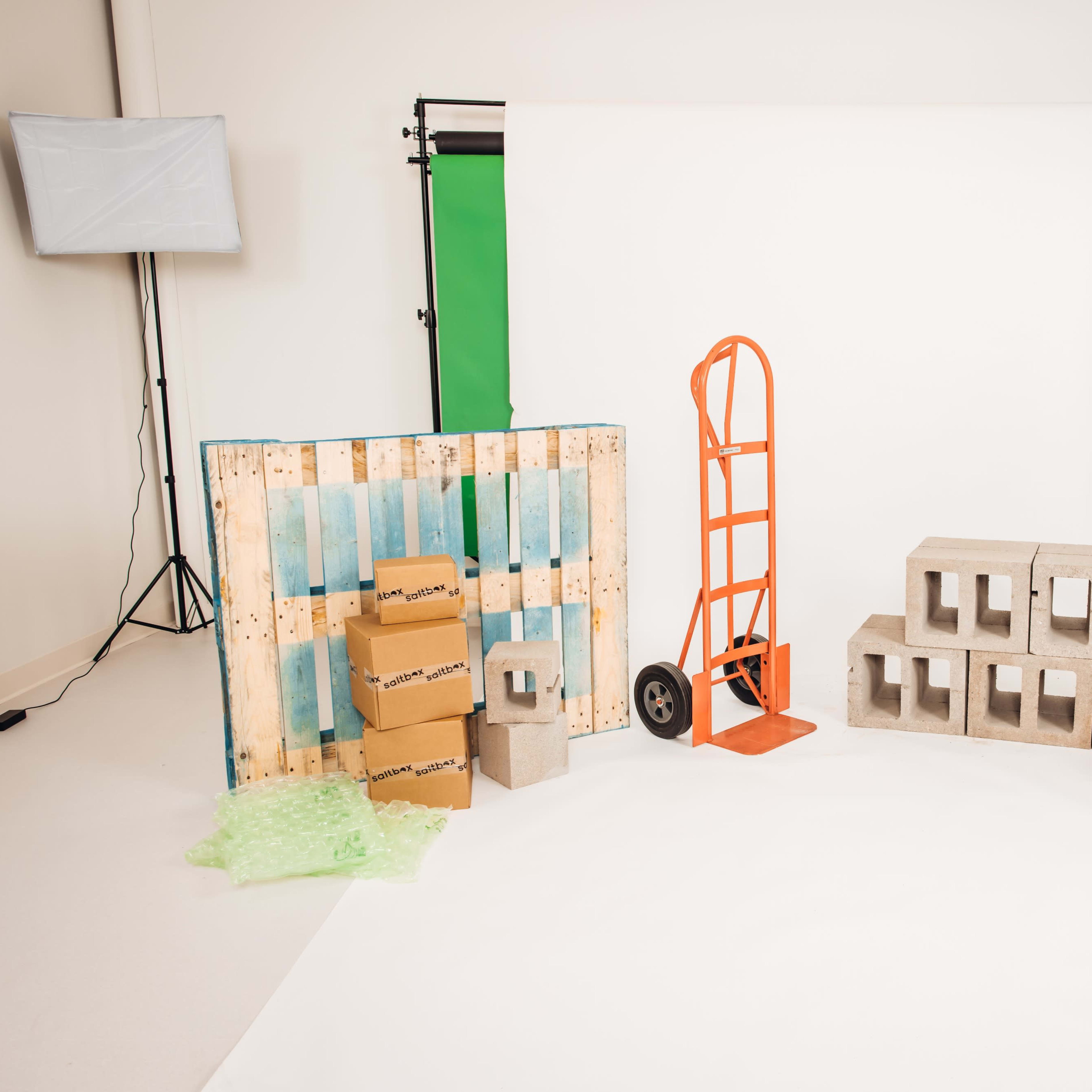 The scene features a studio setup with a green backdrop, several boxes, a hand truck, and concrete blocks arranged on a white floor, illuminated by two soft box lights.