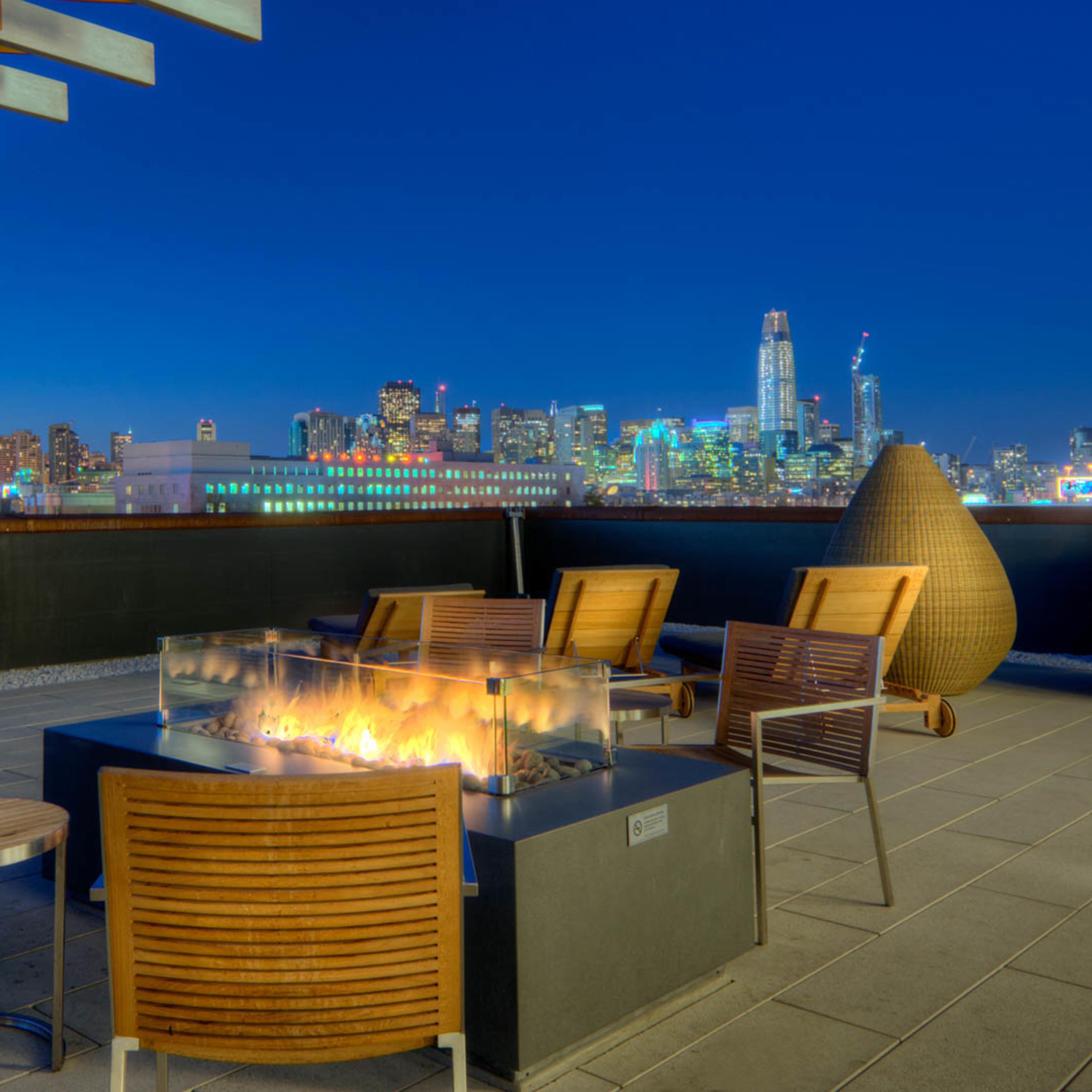 A rooftop terrace features a fire pit surrounded by wooden chairs, with a city skyline illuminated by night in the background.
