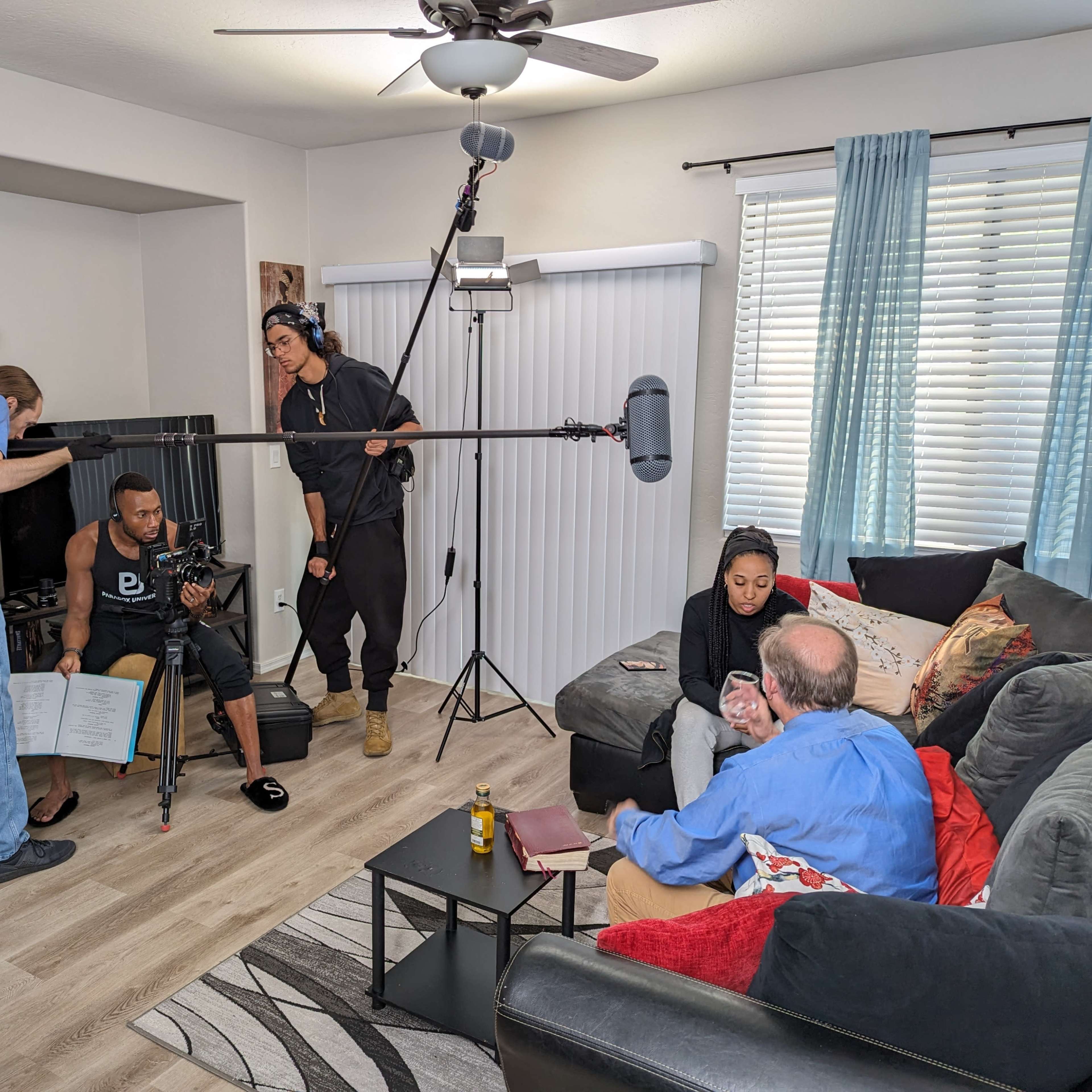 A film crew is recording a scene in a living room, featuring two actors seated on a couch while a sound technician holds a microphone overhead and a camera operator adjusts equipment.