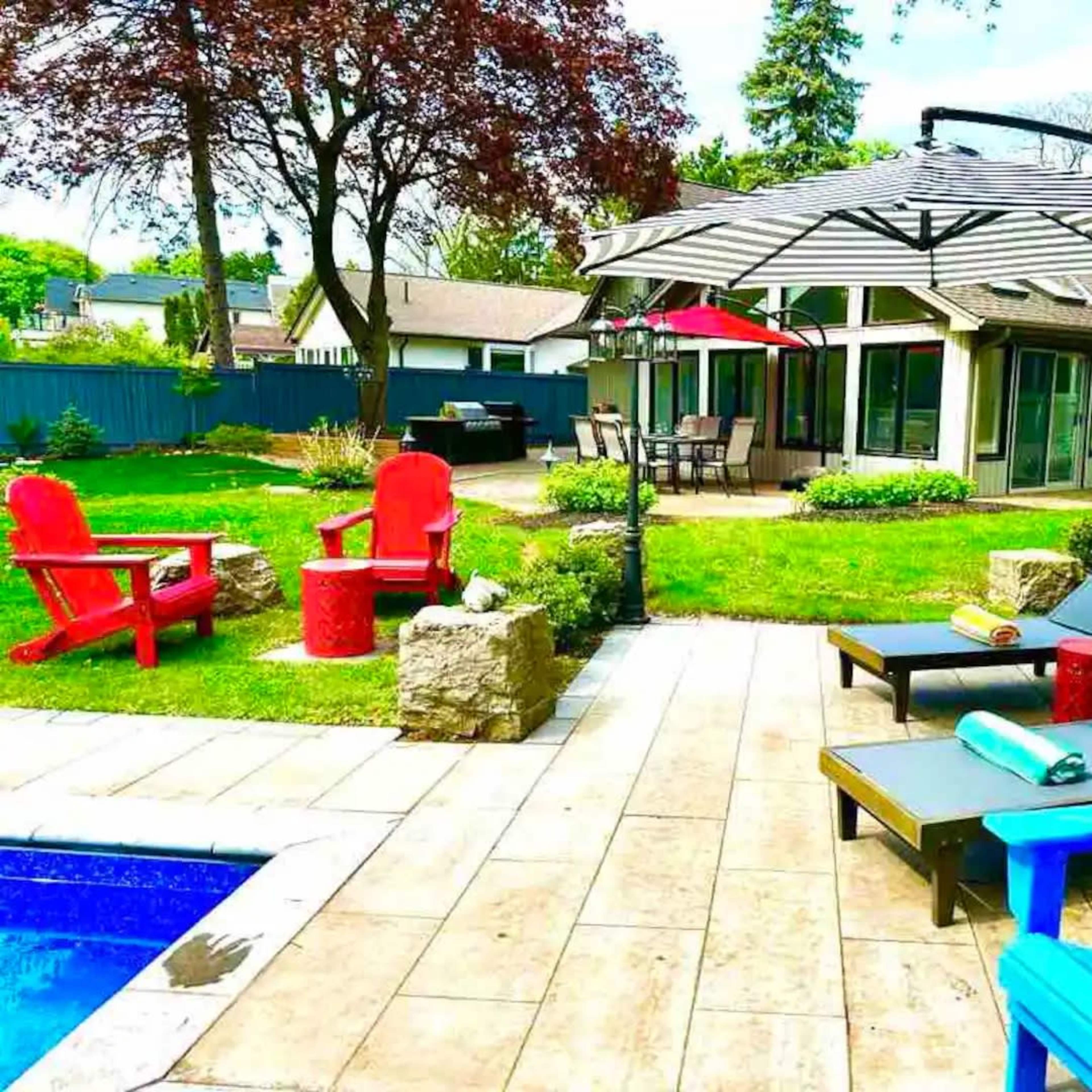 A backyard with a pool, lounge chairs, an umbrella, and a seating area surrounded by greenery and a house in the background.
