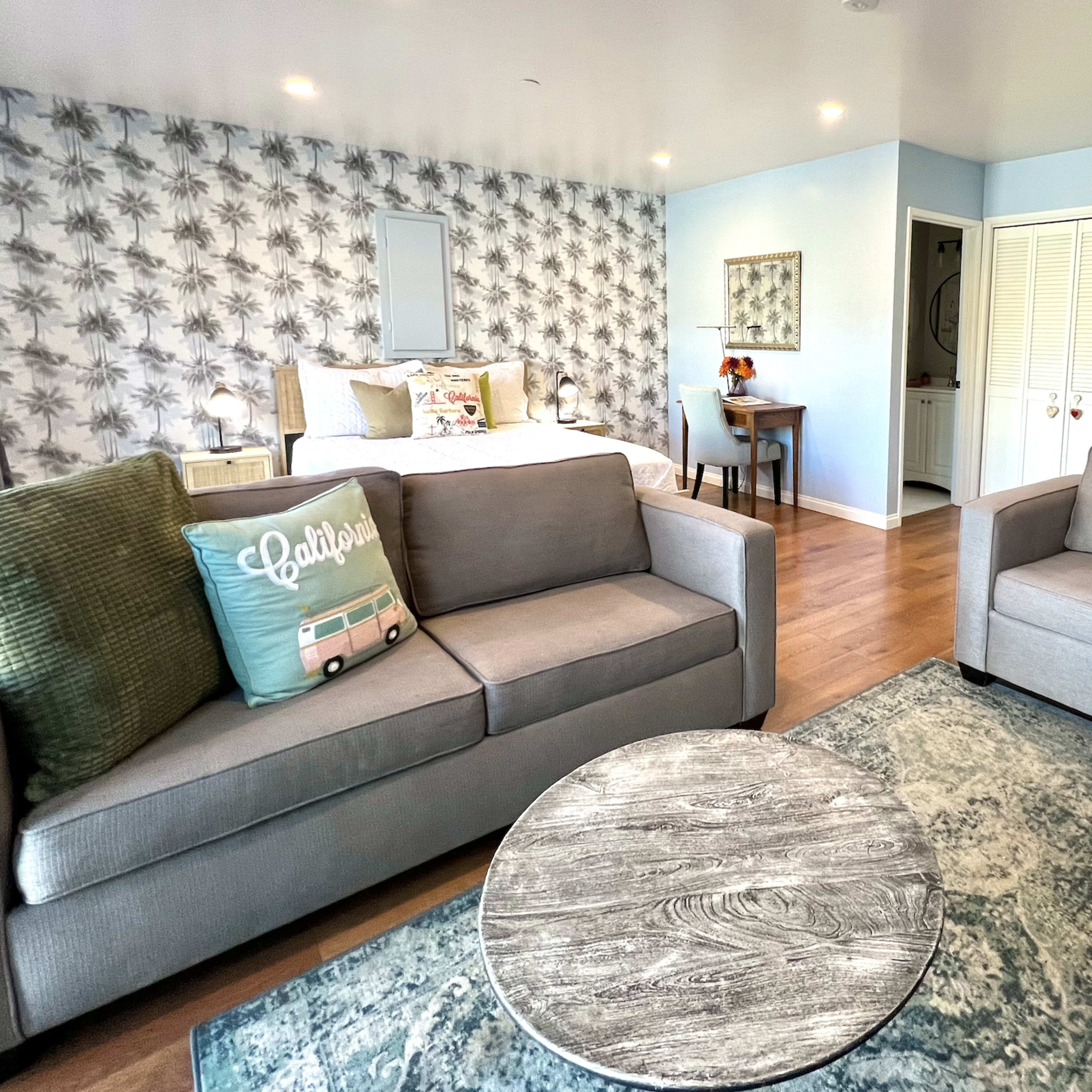 A cozy living space with two gray sofas, a round coffee table, and a bed in the background, all set against a patterned wall and light blue accents.