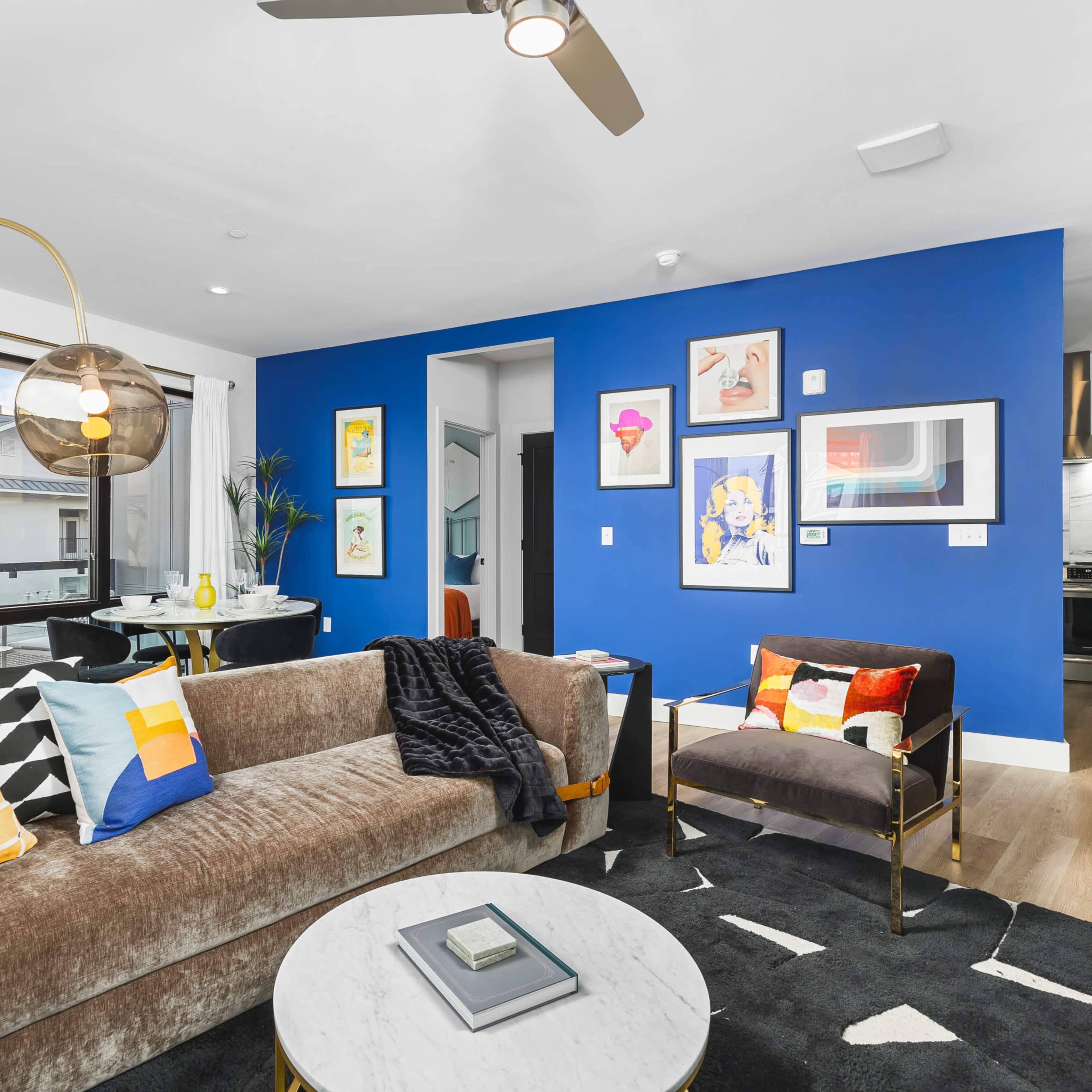 A modern living room featuring a velvet sofa, a round marble coffee table, colorful artwork on blue walls, and a ceiling fan.
