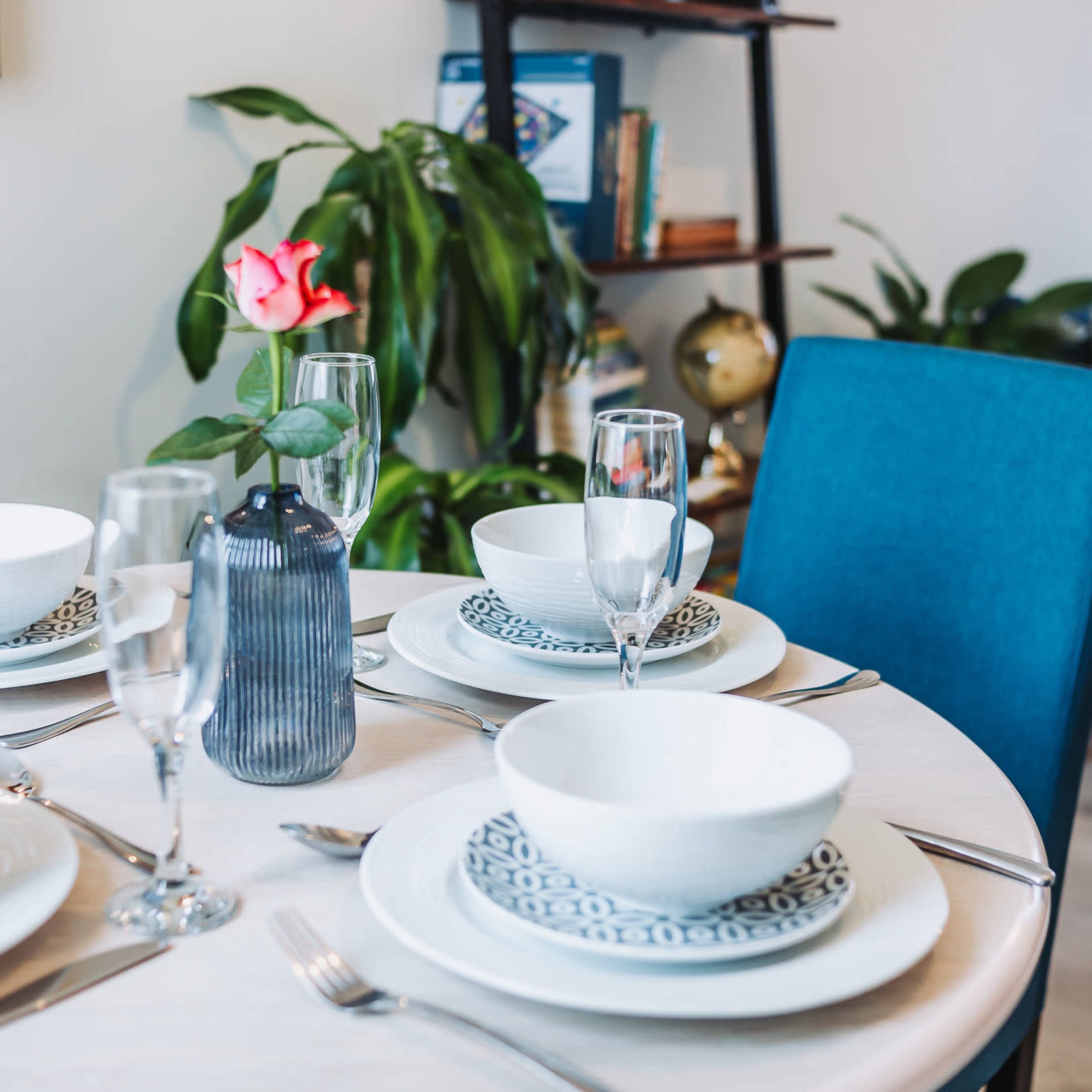 A round dining table is set with white bowls, decorative plates, and glassware, accompanied by a vibrant rose in a vase, against a backdrop of a bookshelf and indoor plants.
