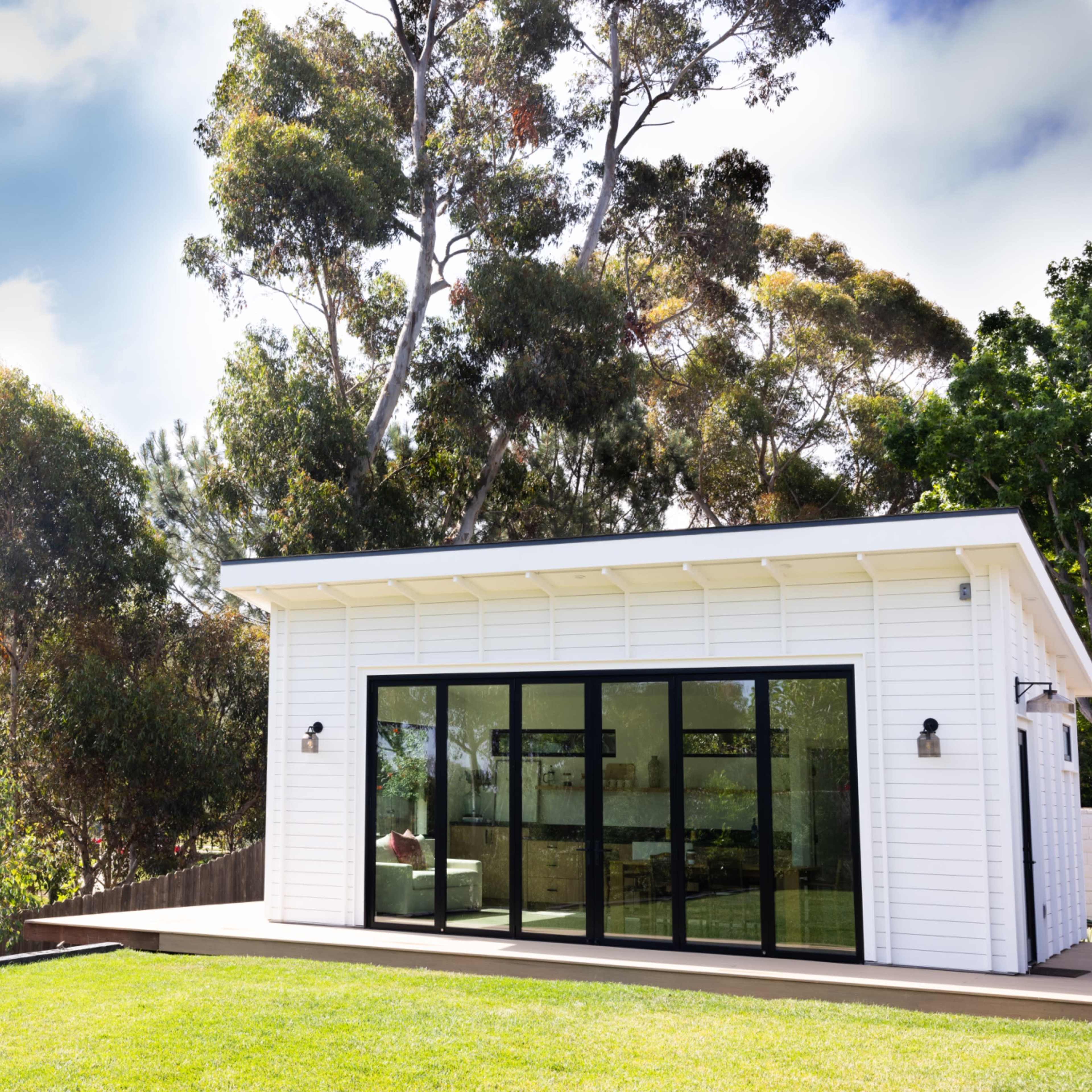A modern white house with large glass doors sits on a grassy lawn surrounded by trees.