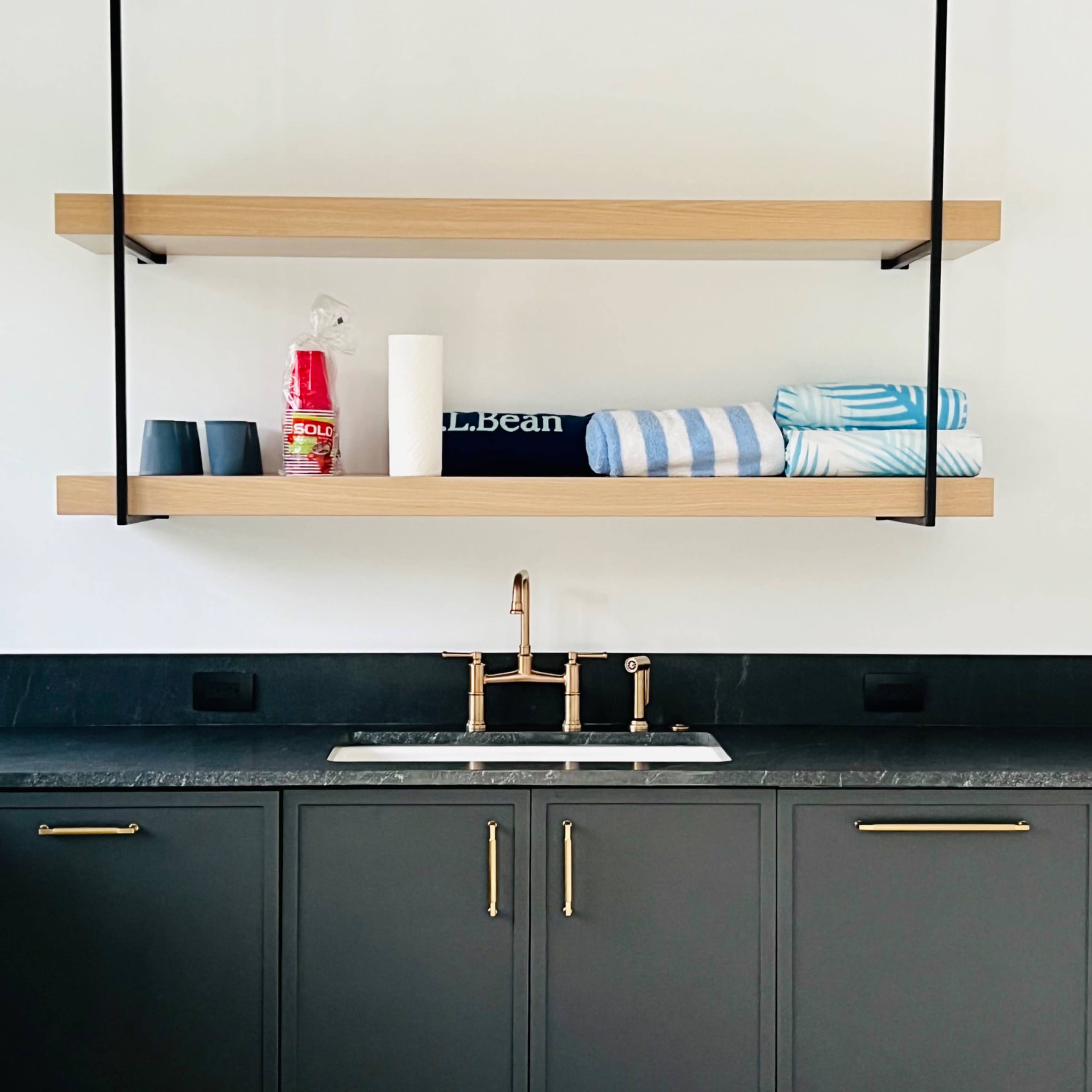 A modern sink area features two wooden shelves above a dark cabinet, displaying neatly folded towels, cups, and cleaning supplies.