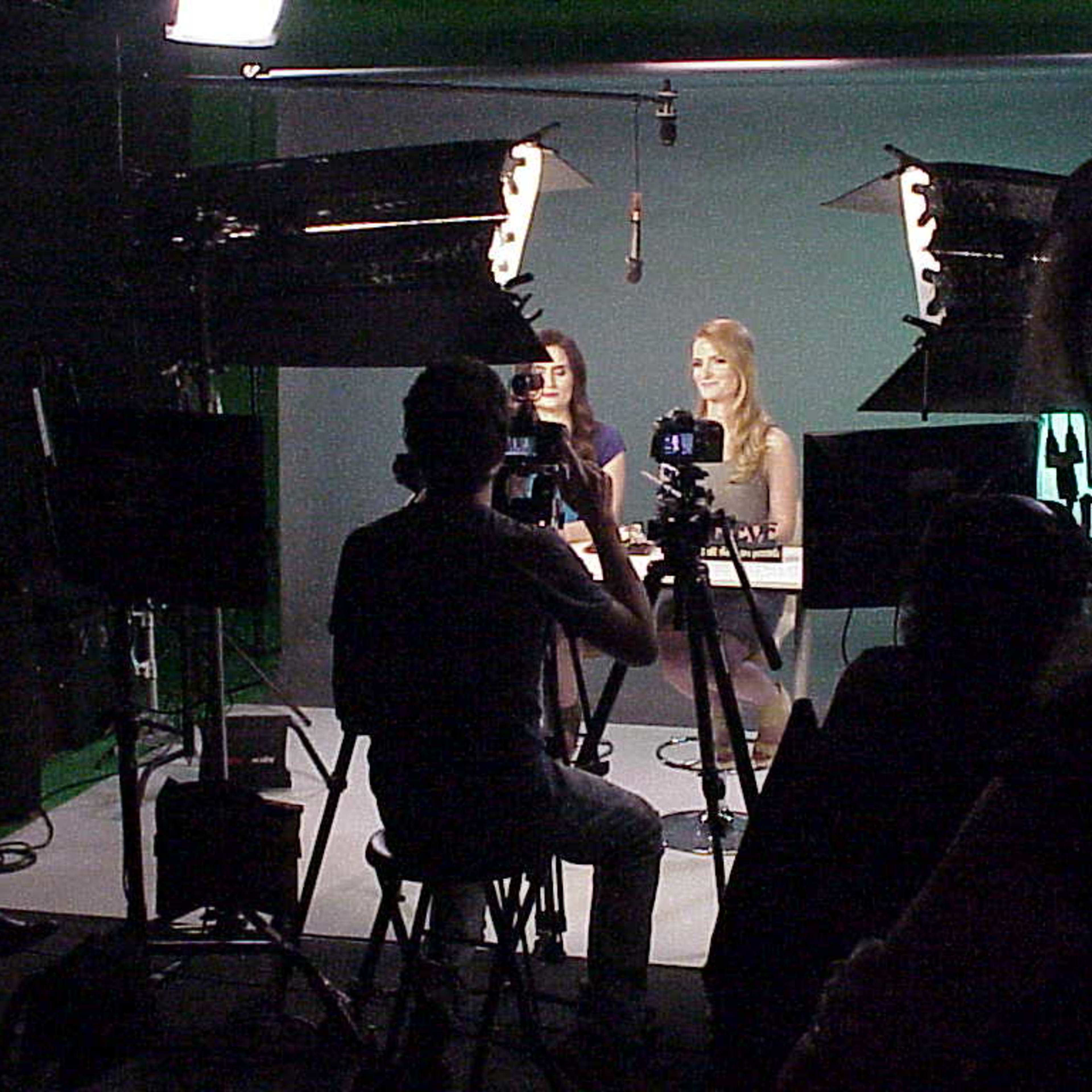 Two women are seated at a table with microphones in front of them, while a person behind a camera captures the scene in a studio setting.