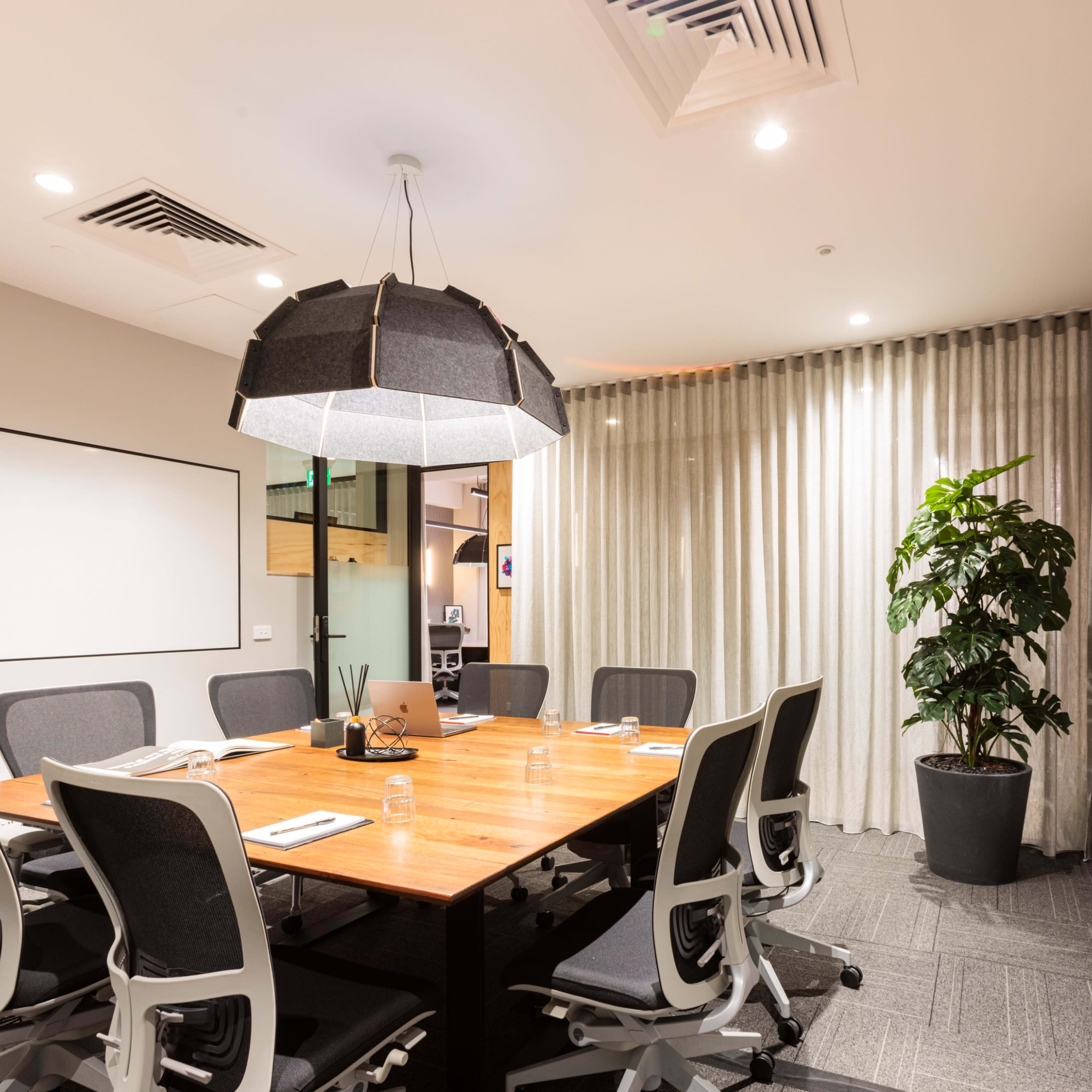 A modern conference room with a large wooden table surrounded by ergonomic chairs, a flat-screen monitor on the wall, and a plant in the corner.