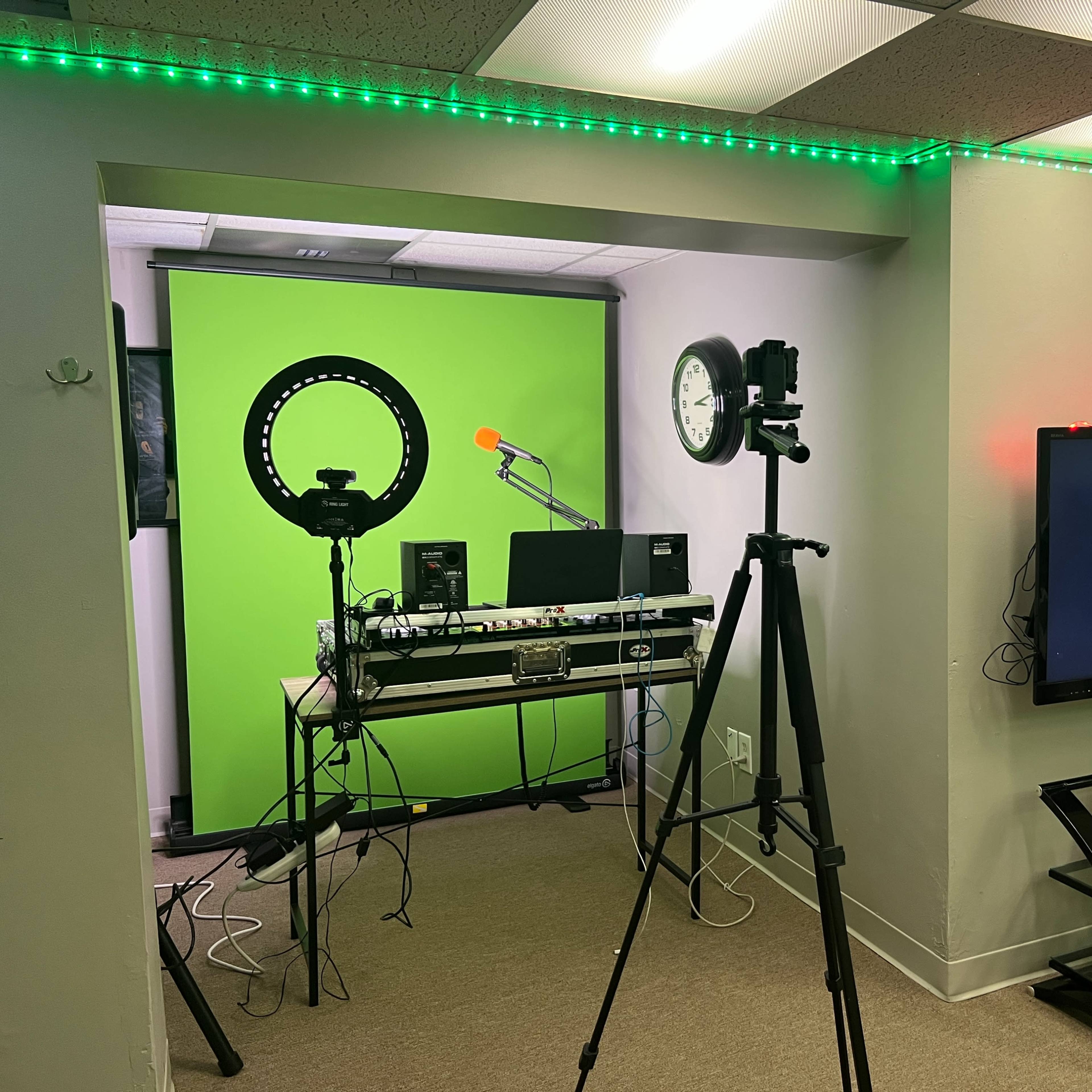 A recording studio setup featuring a green screen, cameras on tripods, a desktop with audio equipment, and a television on a glass stand.