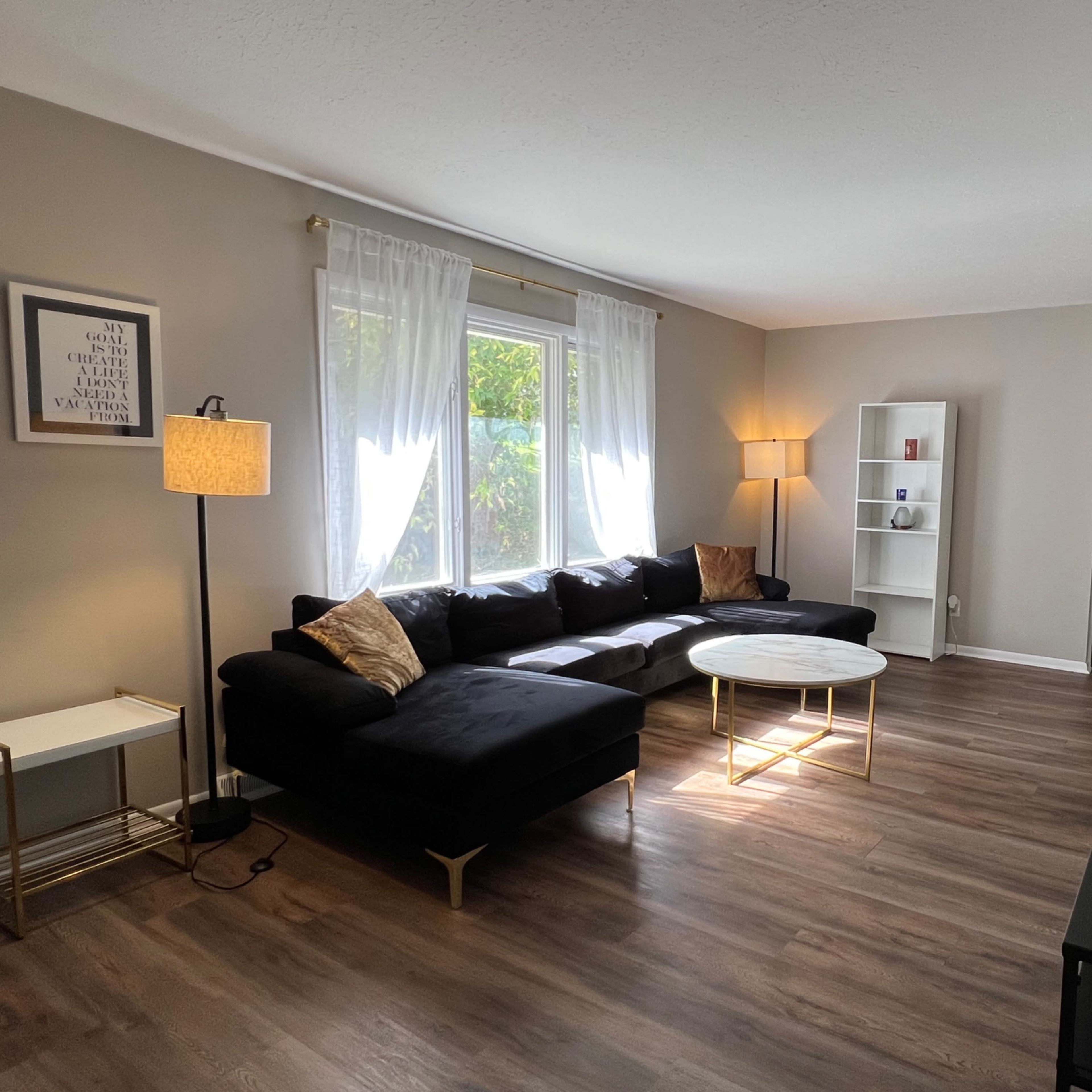 The living room features a dark sectional sofa, a circular coffee table, large windows with sheer curtains, and a floor lamp, all set on hardwood flooring.