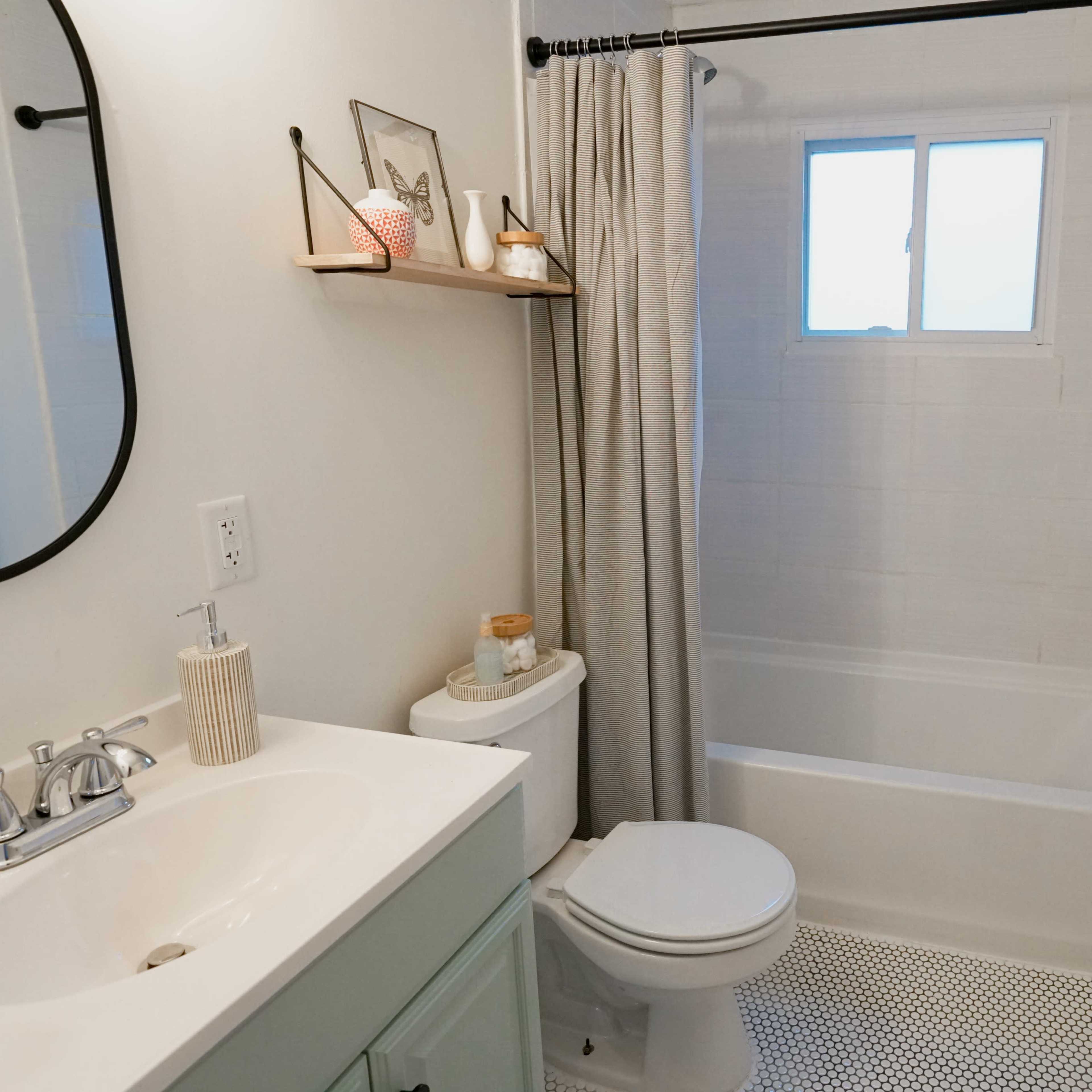 A clean bathroom features a white bathtub, a sink with a mirror, and a window allowing natural light, along with minimal decor on the shelves.