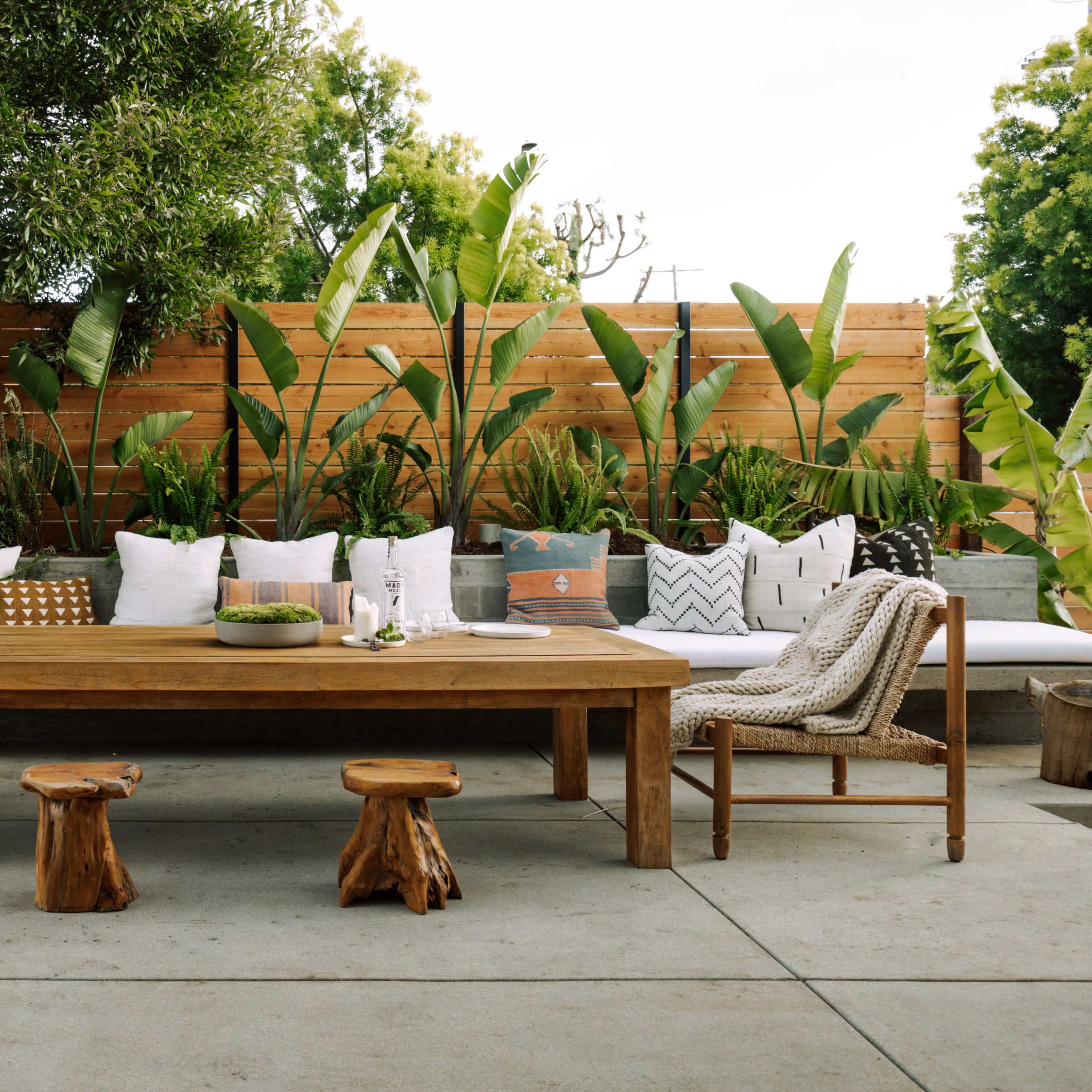 A wooden dining table surrounded by woven chairs and a long bench on a patio, bordered by tropical plants and pillows.
