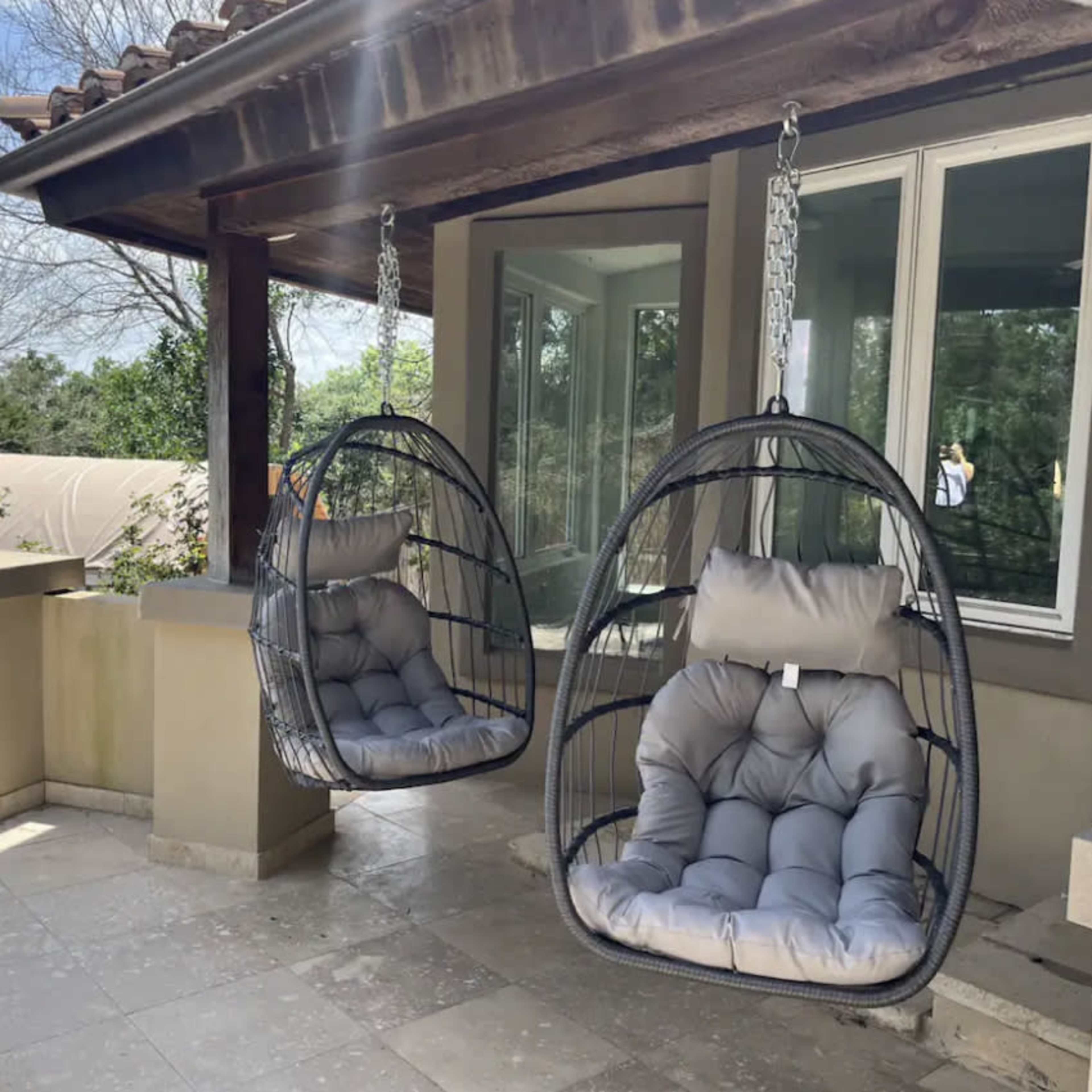 Two hanging wicker chairs with cushions positioned on a porch surrounded by greenery.