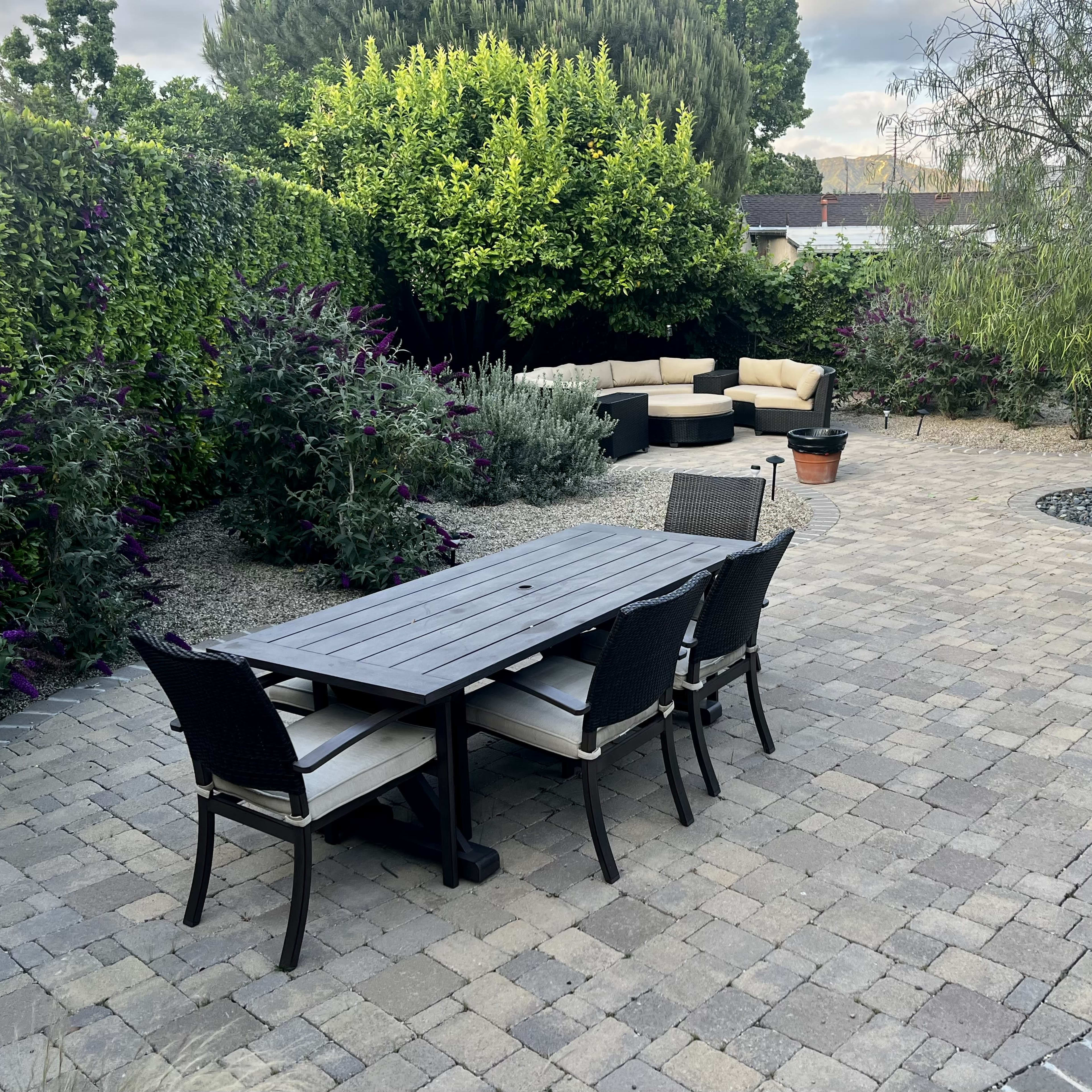 A paved patio area features a rectangular table with chairs, surrounded by garden plants and a sectional seating arrangement.