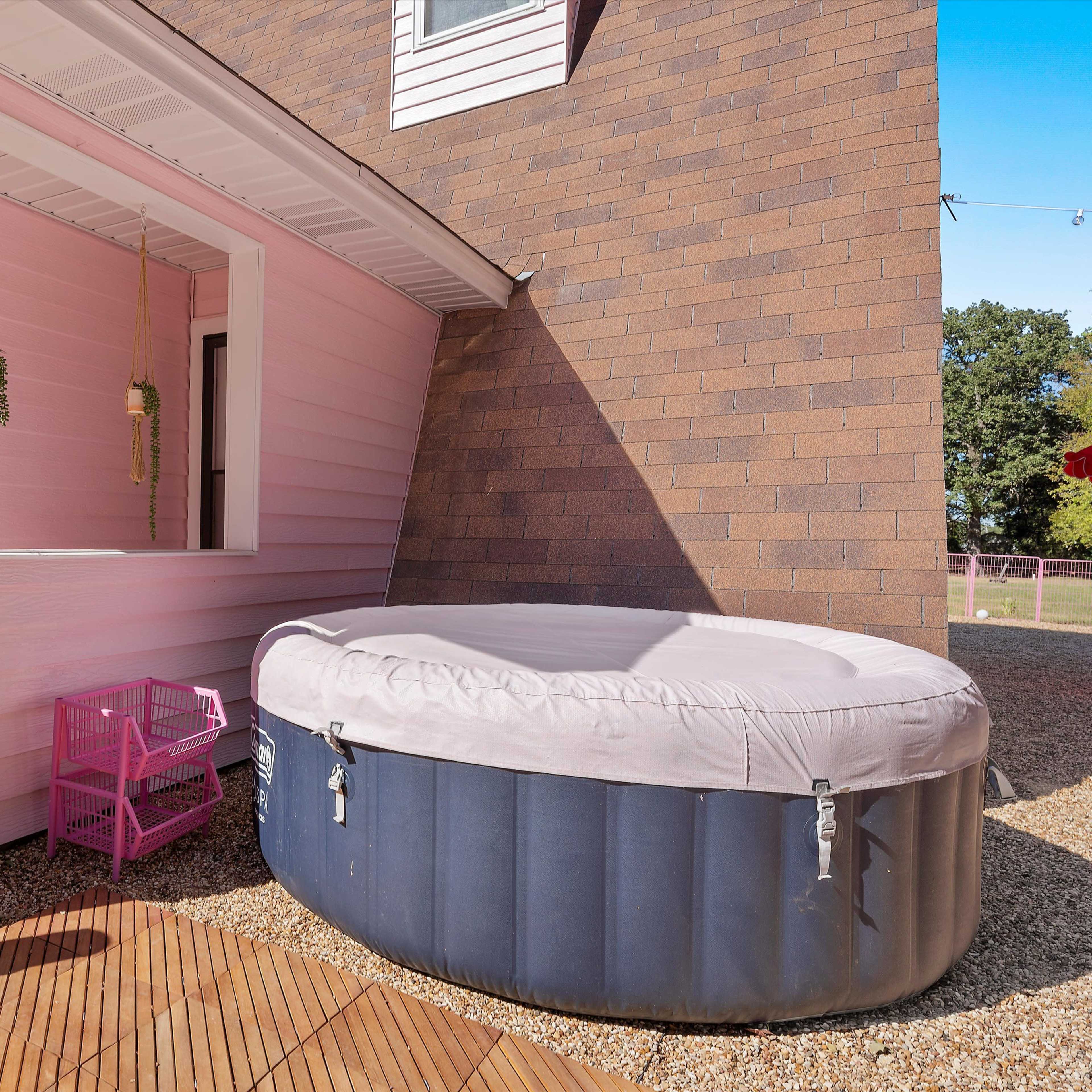 A round hot tub is situated next to a pink wall with a serving window and hanging plants in a gravel area.