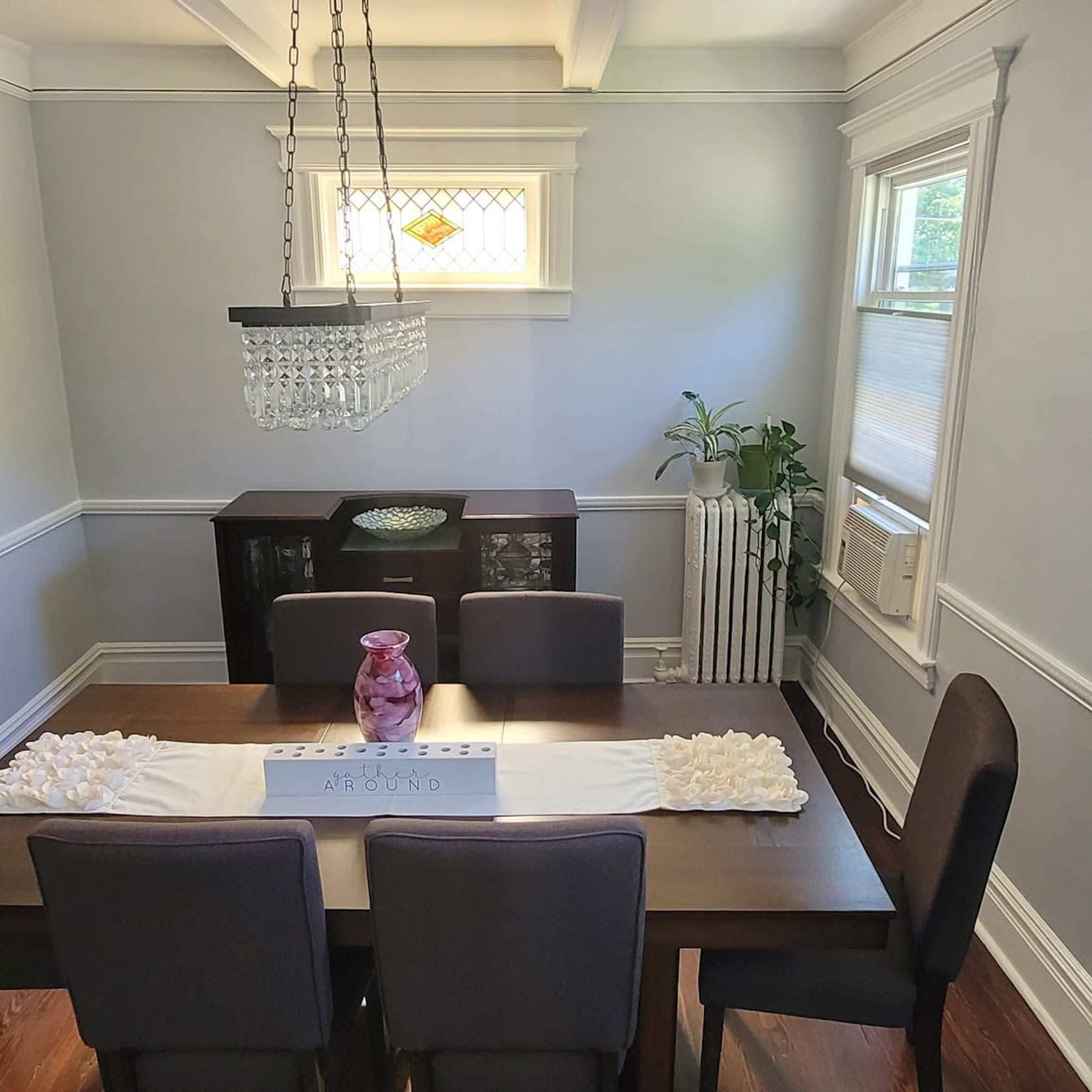 A dining room features a rectangular table with four chairs, a centerpiece, and large windows providing natural light.