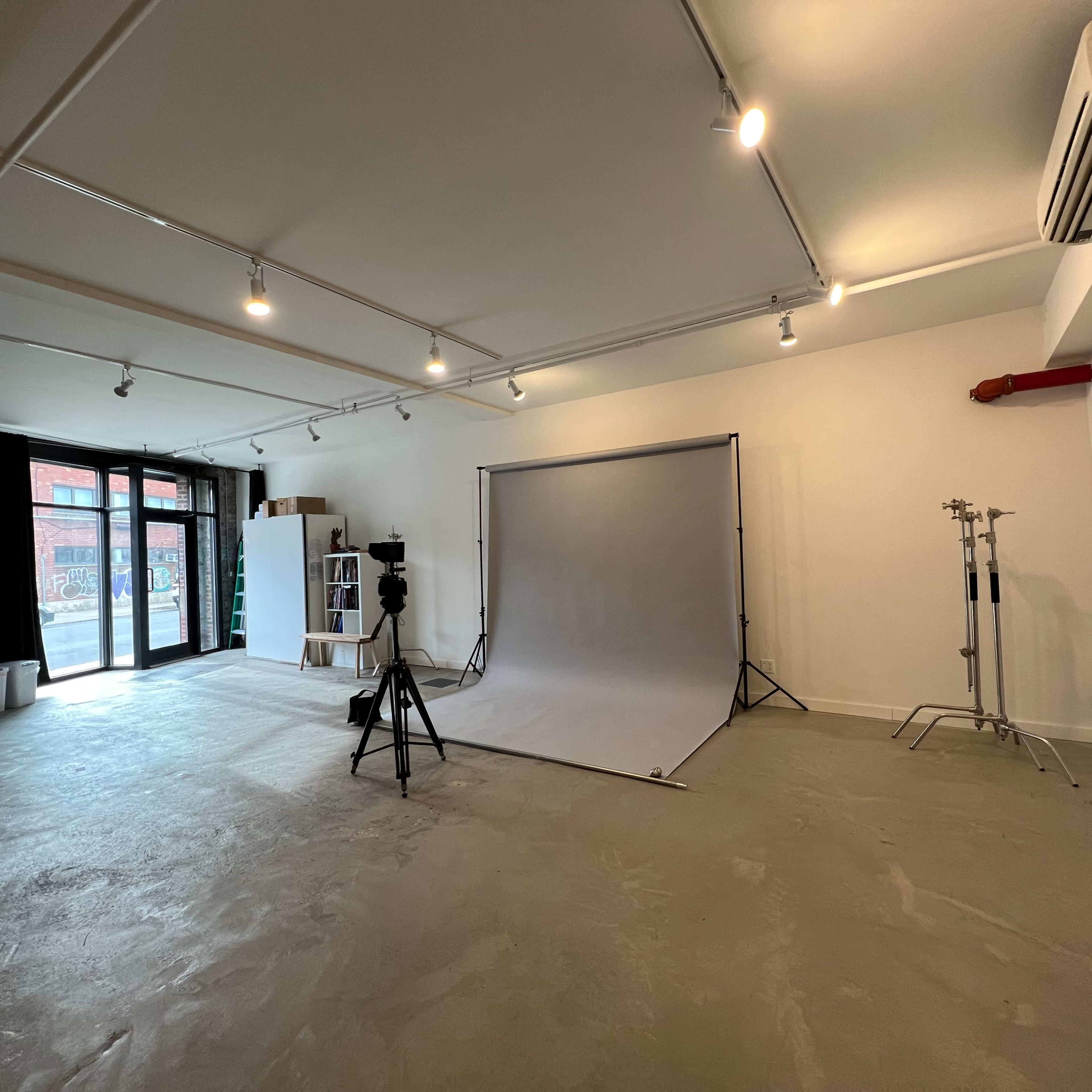 A spacious photography studio with a gray backdrop, lighting equipment, and large windows allowing natural light to enter.
