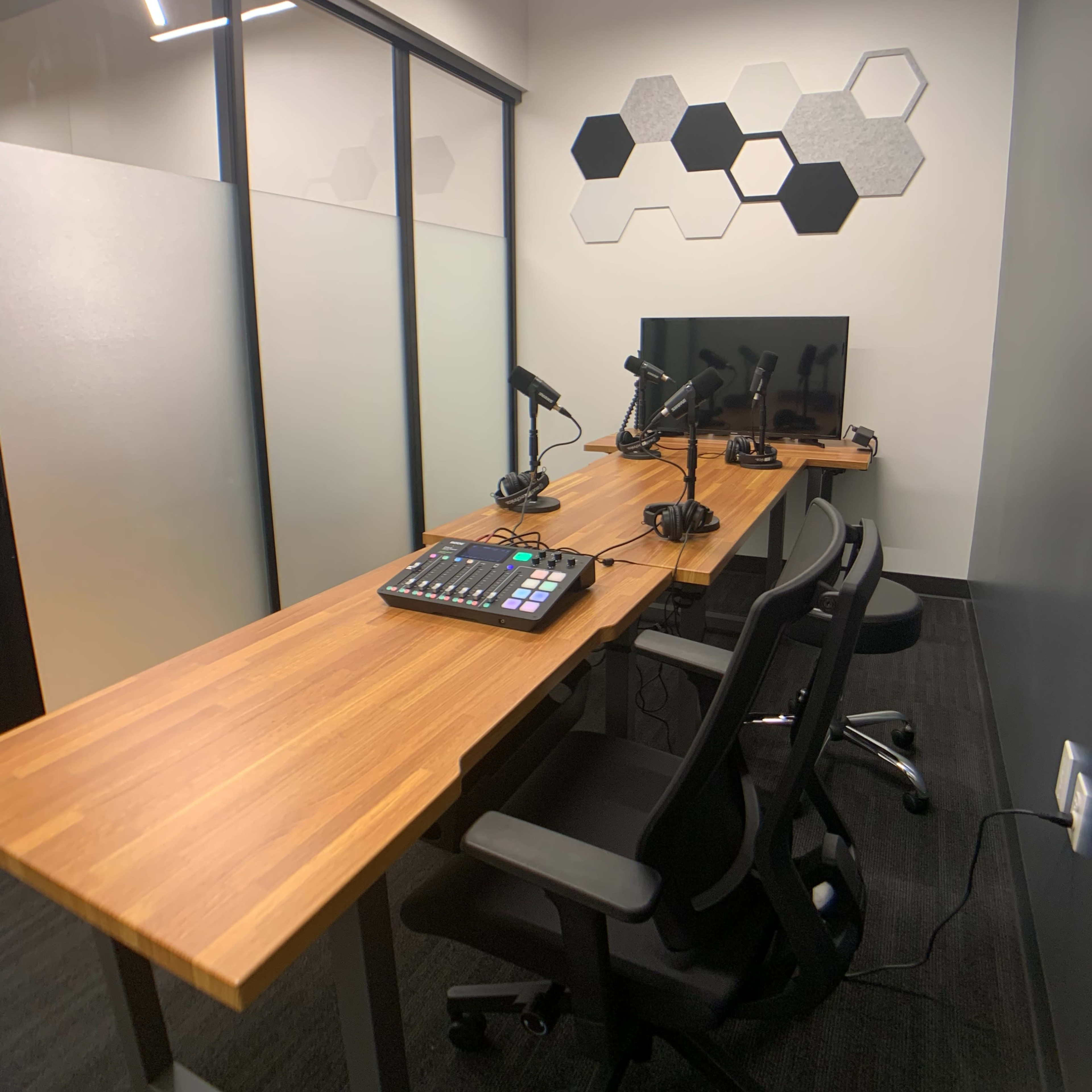 A small, modern workspace with a wooden tabletop, two chairs, a computer monitor, and multiple microphones arranged for recording or broadcasting.