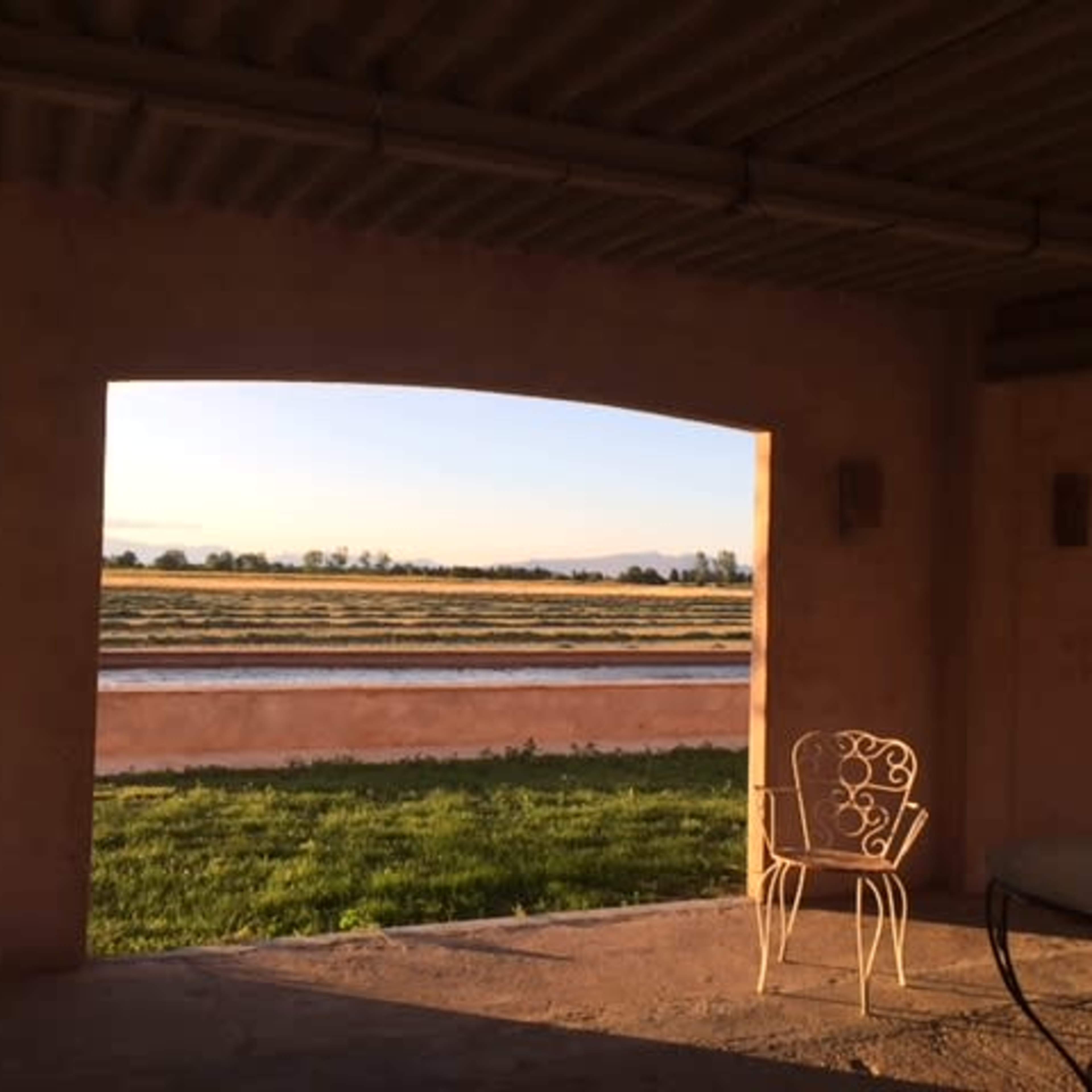 A patio area with a metal chair and a nearby table, overlooking a grassy field and distant horizon under a clear sky.