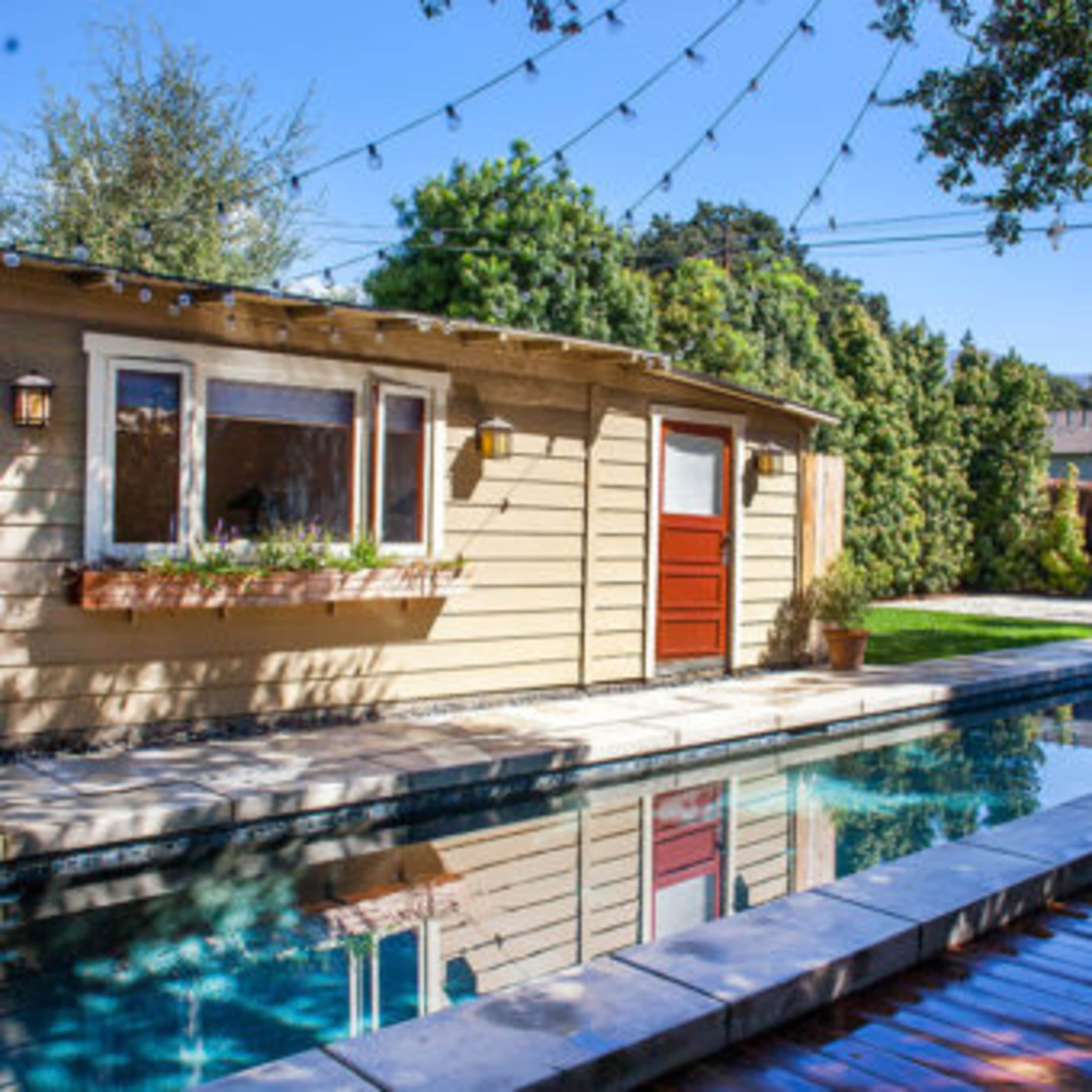 A backyard with a swimming pool, a small wooden shed, and string lights overhead, surrounded by trees and landscaped greenery.