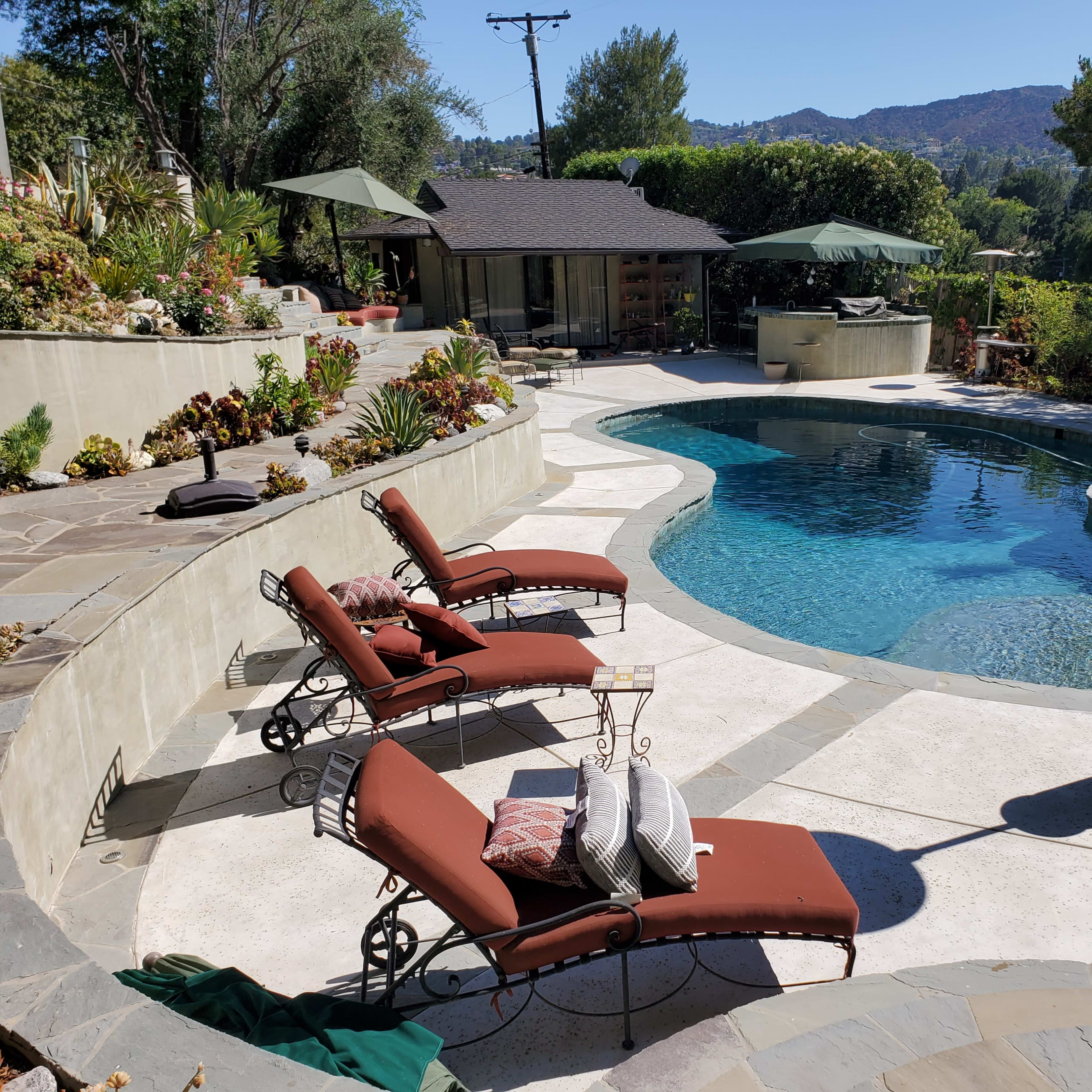 A landscaped backyard with a swimming pool, two lounge chairs, a spa, and several umbrellas surrounded by greenery and mountains in the background.