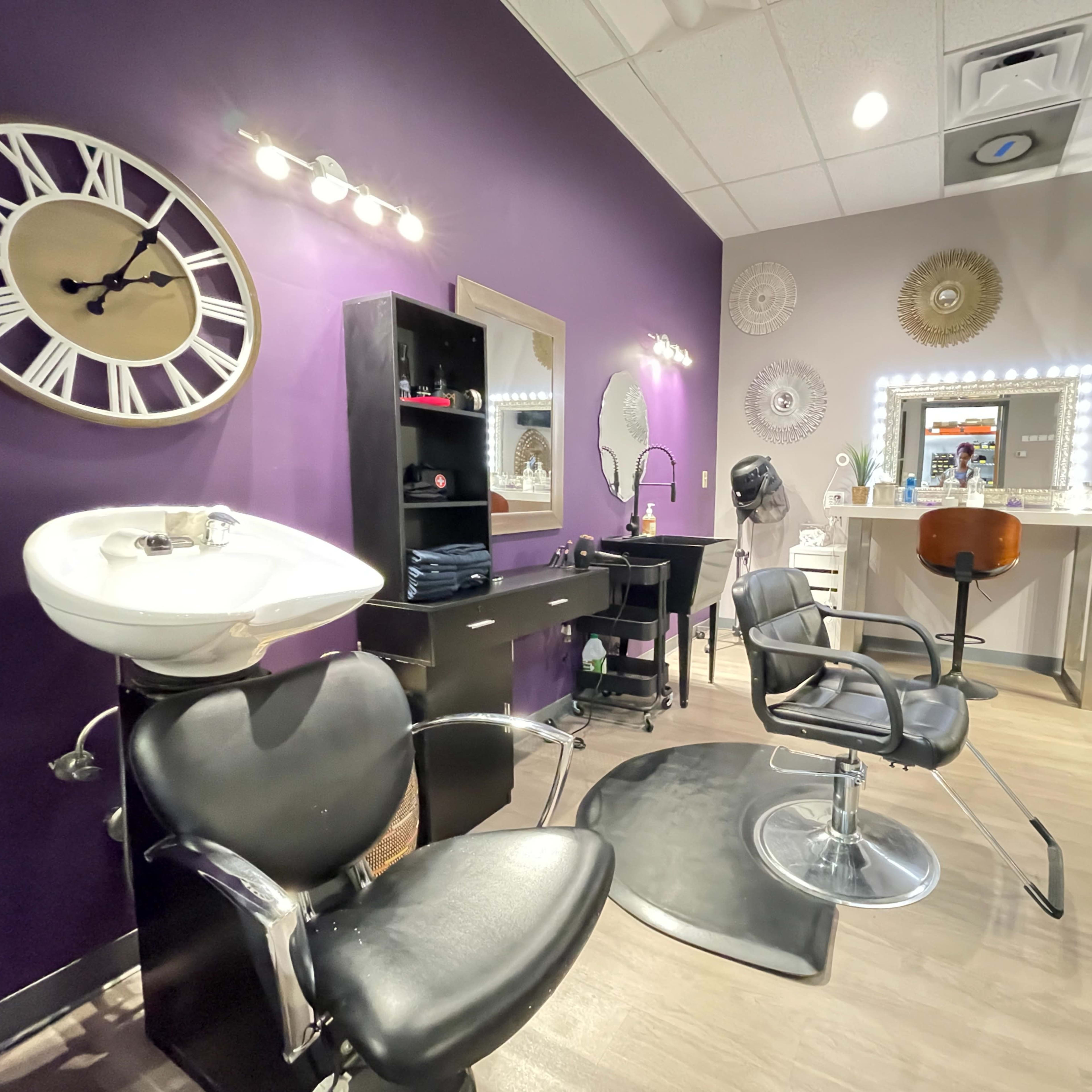 A modern salon interior featuring black styling chairs, a wash station, a large mirror, and decorative wall ornaments, all set against a purple wall.