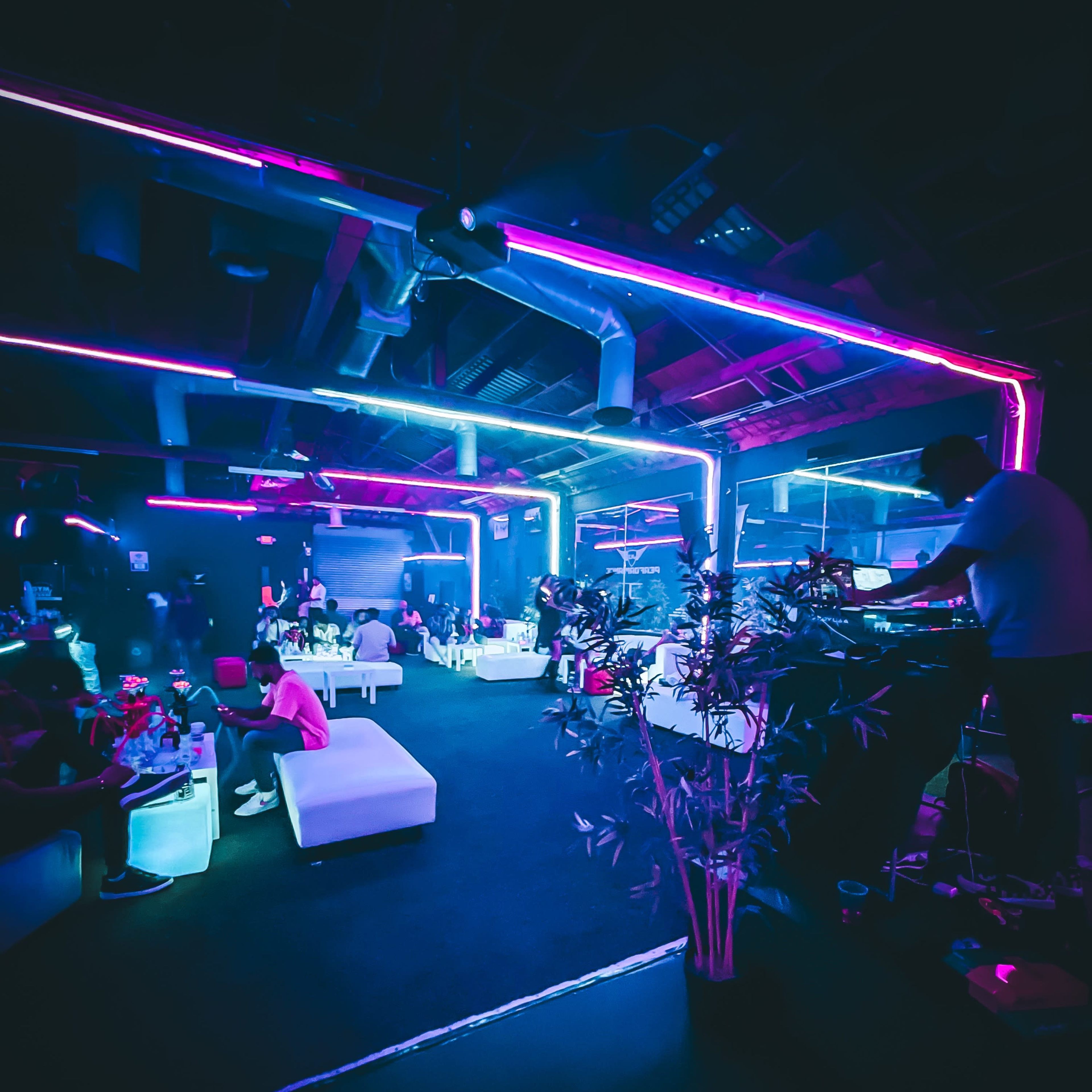 A dimly lit lounge with neon lights, featuring white seating areas and a DJ setup in the corner.