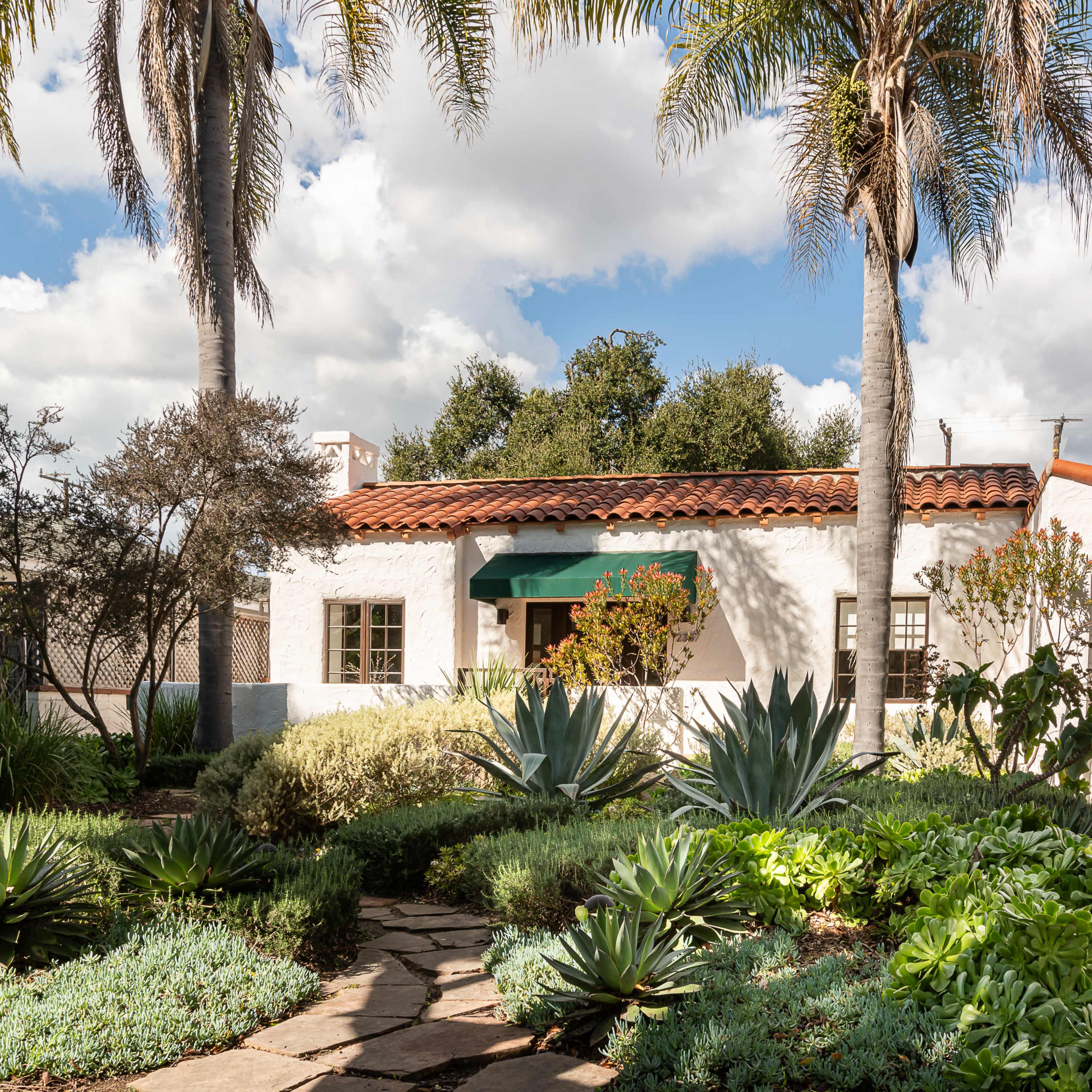 A single-story white house with a red tile roof is surrounded by various plants and a stone pathway, featuring a garage on the right.
