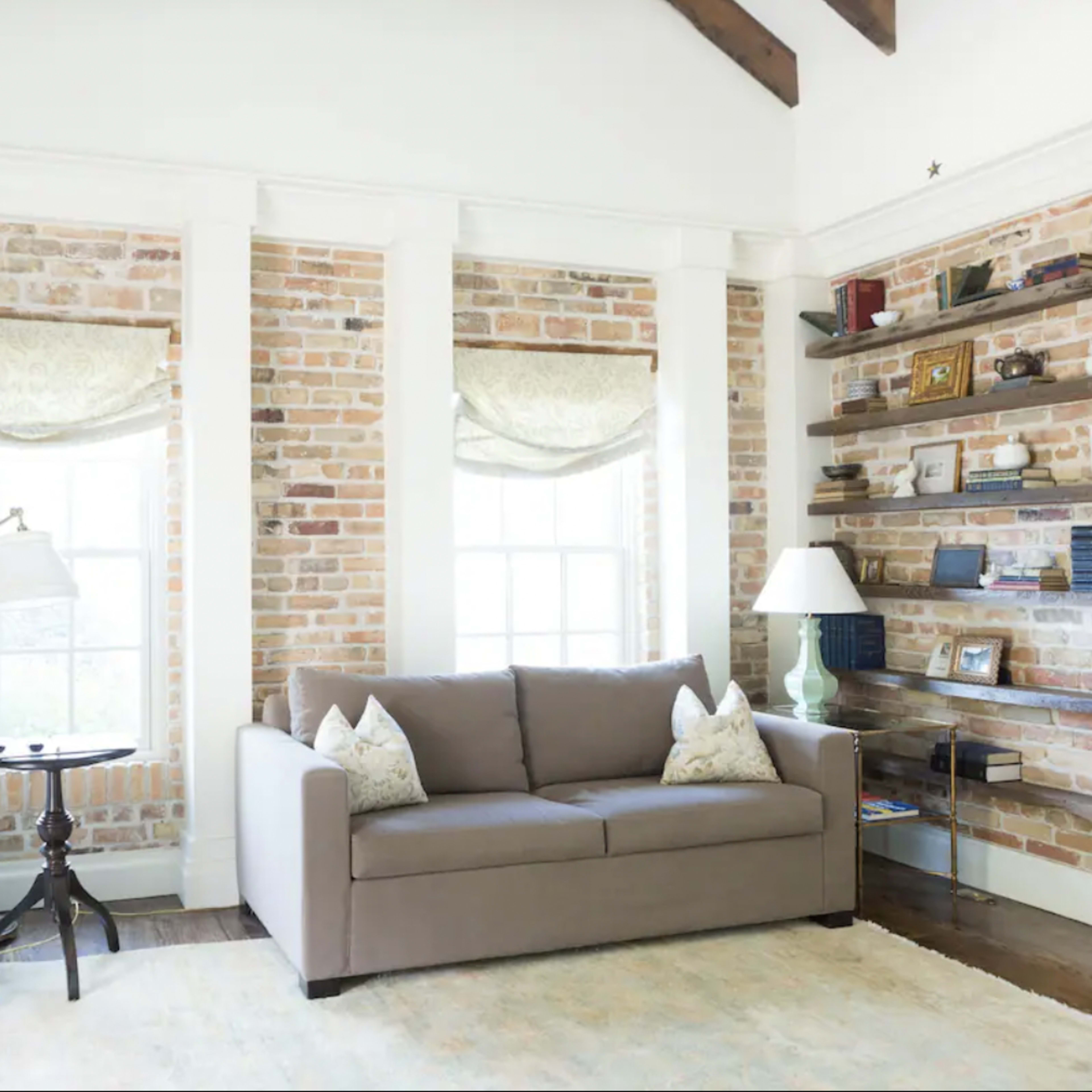 A living room with exposed brick walls, a light-colored sofa, an armchair, a side table, and bookshelves filled with various items.