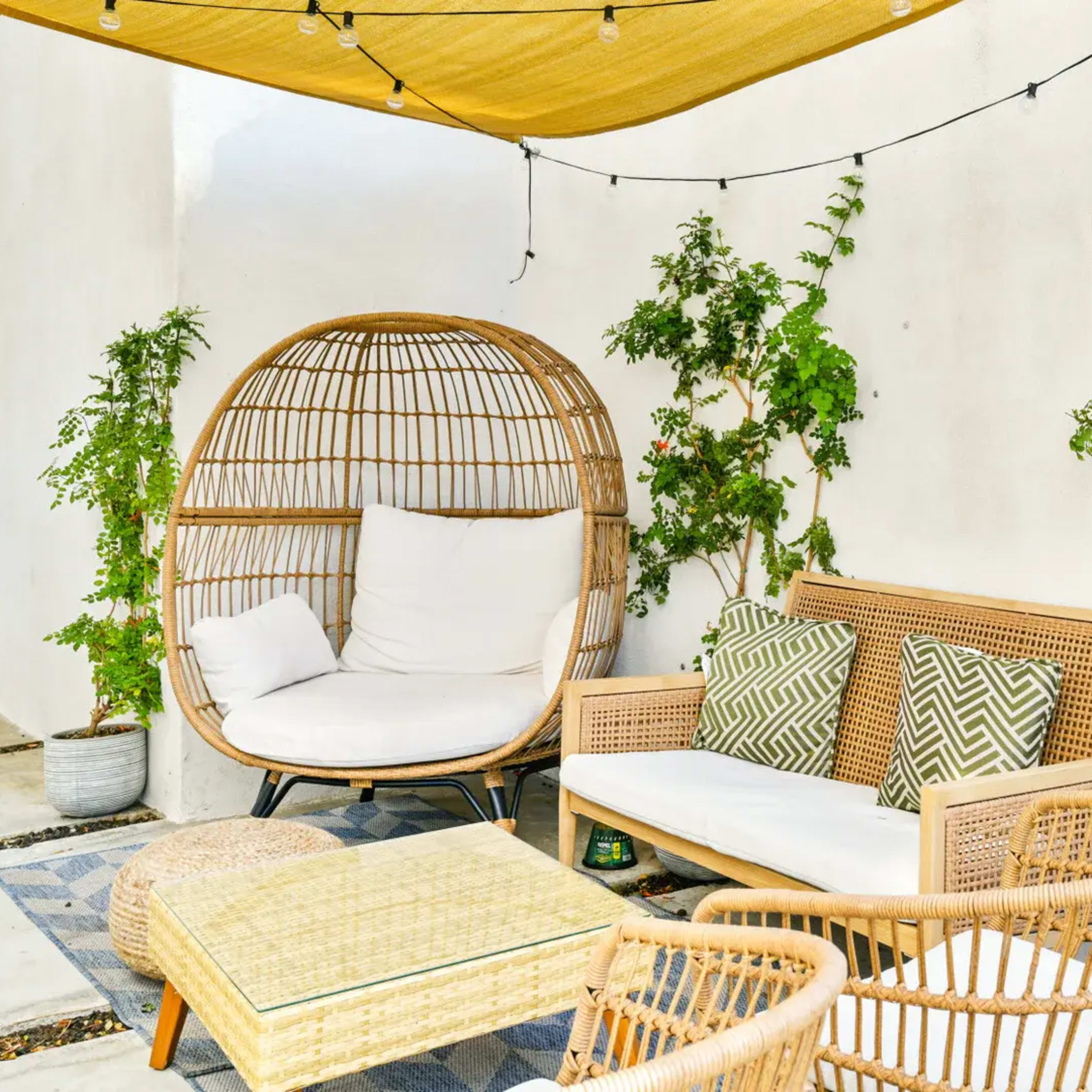 The scene features a cozy outdoor seating area with a circular rattan chair, a cushioned sofa, and potted plants against a white wall, all under a yellow canopy and string lights.